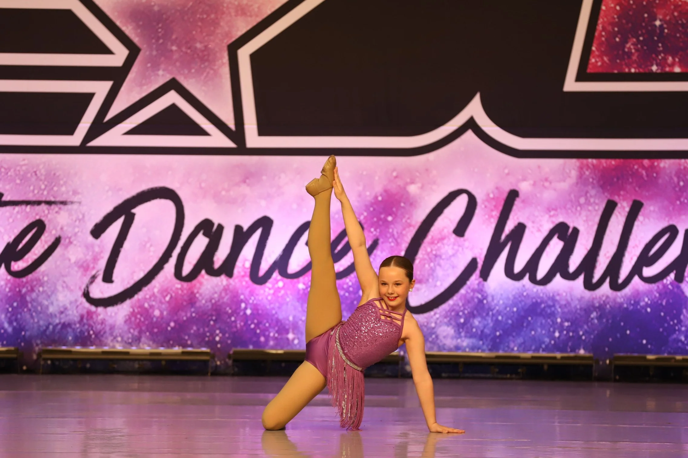 Young girl in purple costume performing a split on stage at a dance competition with colorful galaxy-themed backdrop reading "Little Dance Challenge."