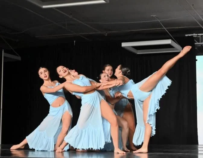 Six dancers in matching light blue costumes performing a choreographed dance on stage with black curtains in the background.