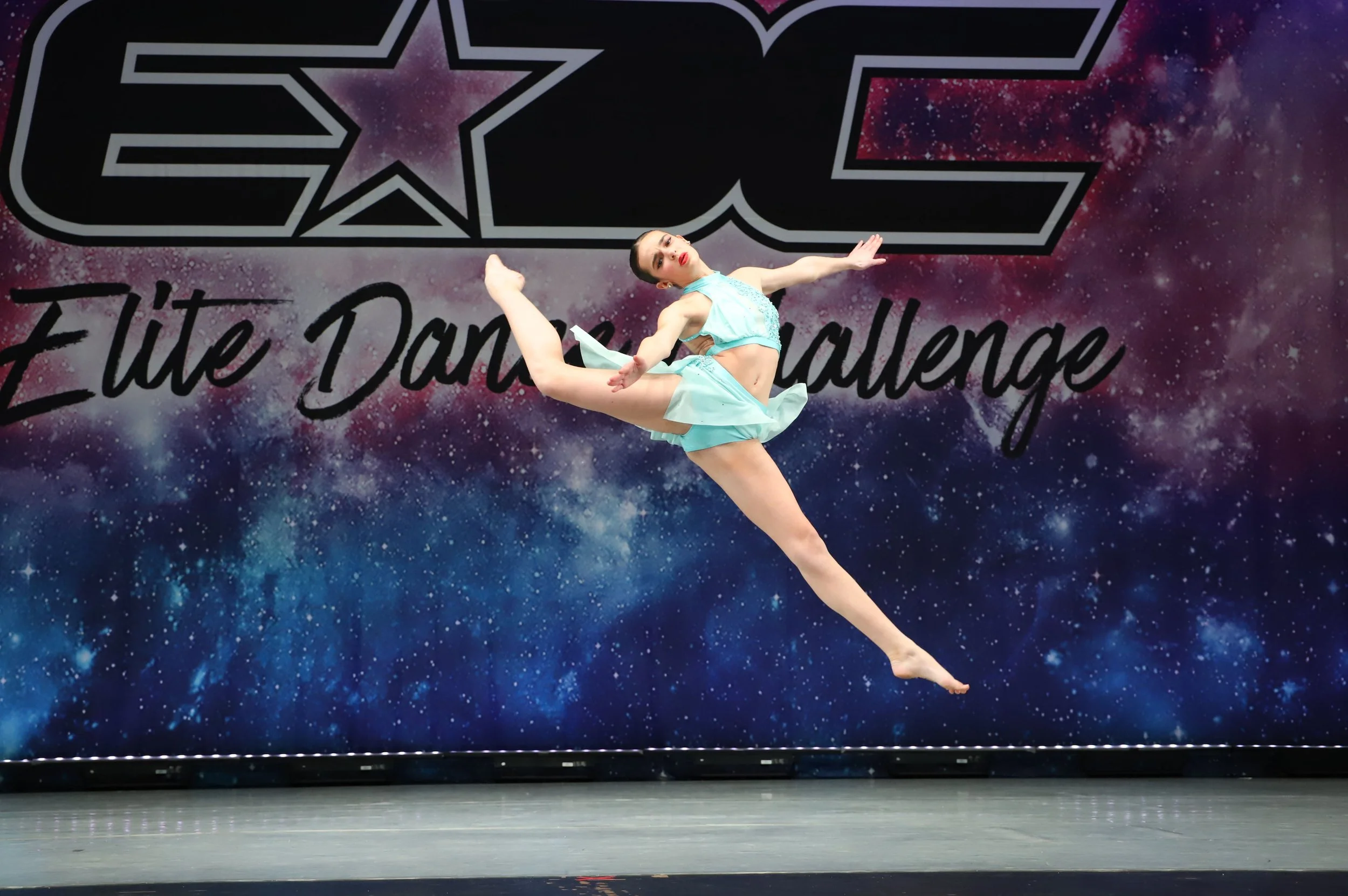 A female gymnast in a light blue costume performing a dance move on stage with a galactic space background and the word 'ECCE' in large letters.
