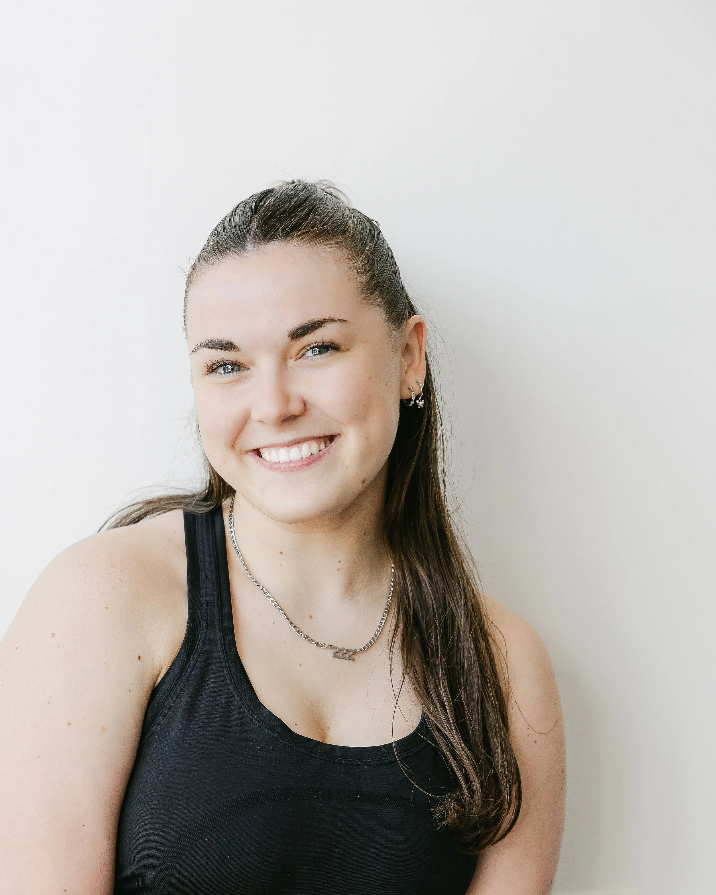 Close-up of a young woman with long brown hair wearing a black tank top, silver jewelry, blue eyes, and smiling, against a plain white wall.