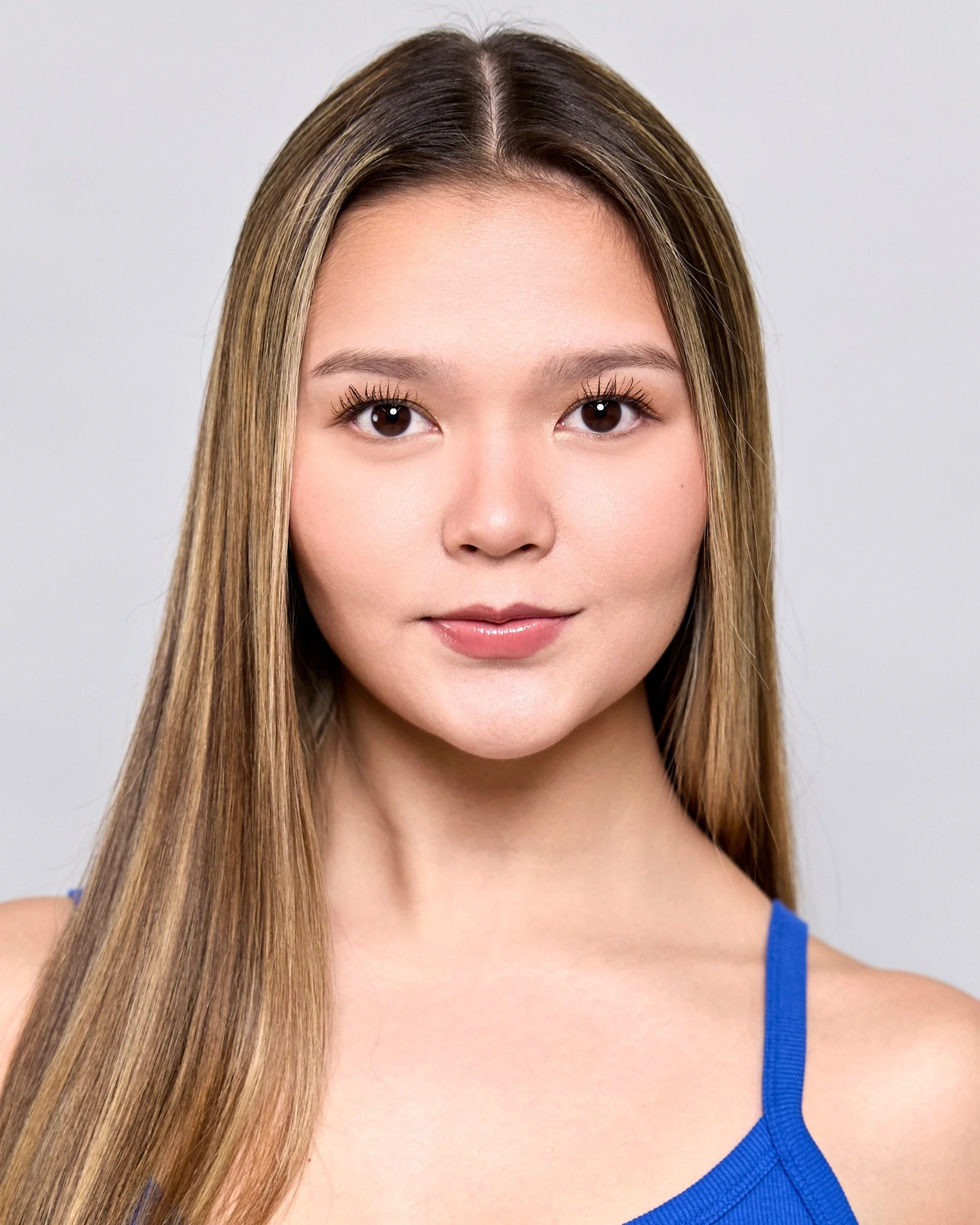 Close-up of a young woman's face with smooth skin, brown eyes, and long straight hair. She is wearing a blue top with thin straps, and the background is plain gray.