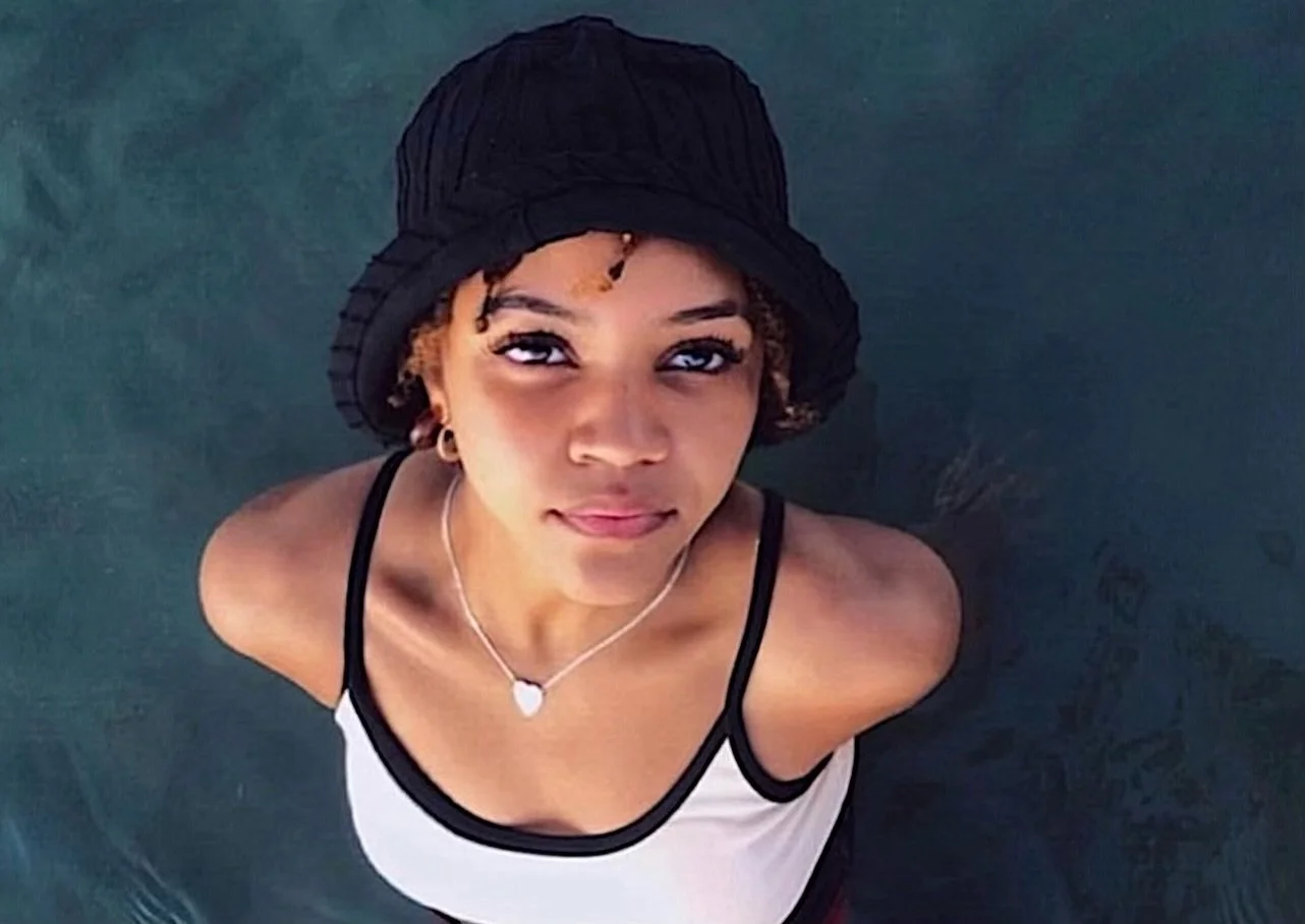 Young woman in a black hat and sleeveless top standing in water, looking up at the camera.