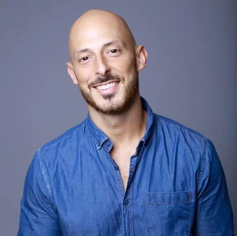 A bald man with a beard smiling, wearing a blue collared shirt, against a plain gray background.