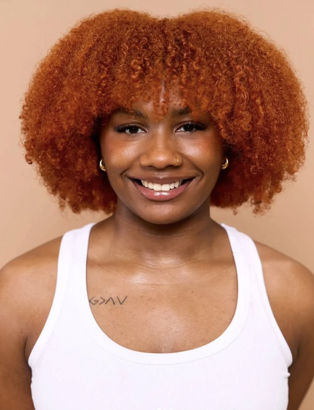 A woman with a big, curly, red hair smiling, wearing a white tank top, gold hoop earrings, and a nose ring, against a beige background.