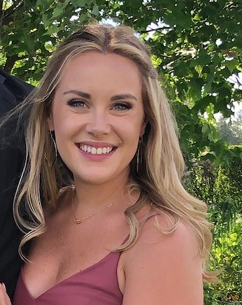 A smiling young woman with blonde hair and blue eyes, wearing a pink sleeveless top, outdoors with green trees in the background.