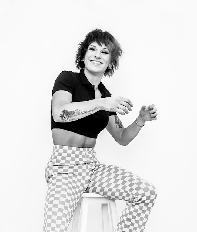 Black and white photo of a woman with short wavy hair, smiling, sitting on a stool, wearing a cropped black shirt and checkered pants.