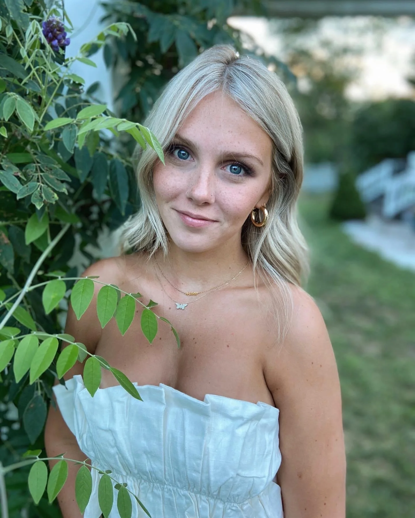 A woman with blonde hair and blue eyes wearing gold hoop earrings and a delicate necklace, standing outside near green foliage, smiling softly.