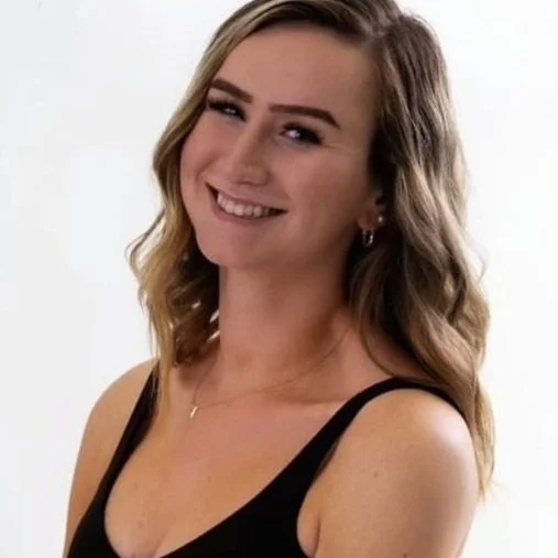 Young woman with wavy blonde hair smiling, wearing a black tank top and a small necklace, against a plain white background.