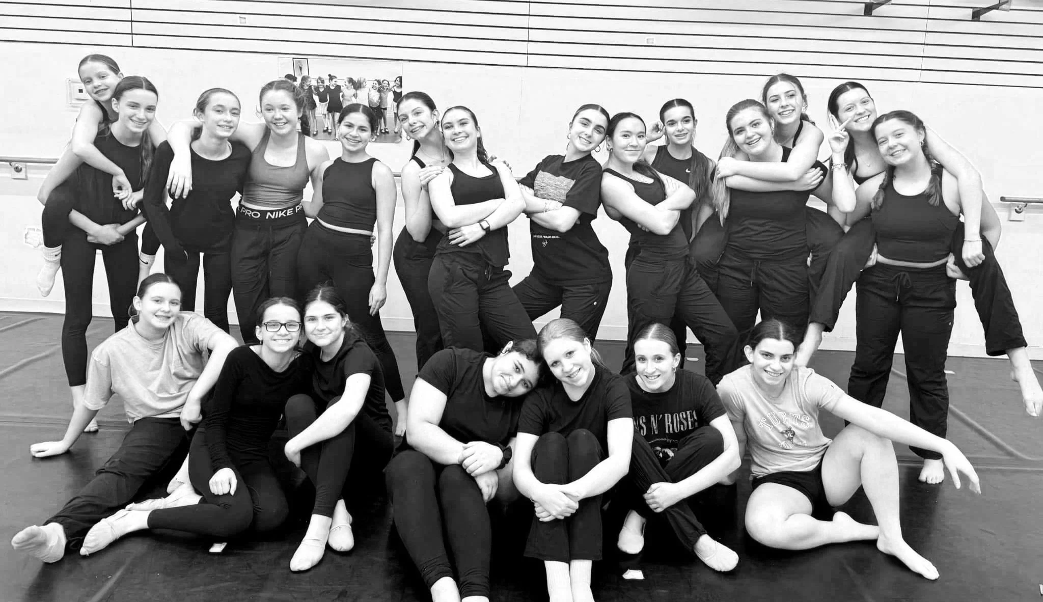 Group of young female dancers in dance studio, some standing and some sitting on the floor, smiling at the camera.