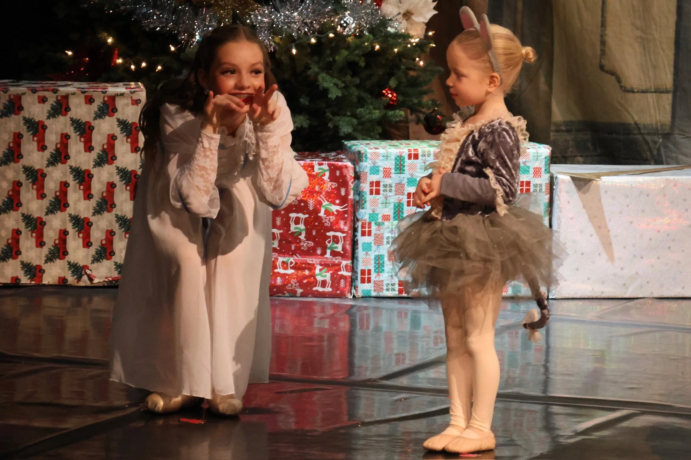 Two young girls, one dressed as a princess and the other as a bunny, are on stage in front of a decorated Christmas tree with wrapped presents underneath. The girl dressed as a princess has curly hair and is wearing a light-colored dress, while the girl dressed as a bunny has her hair pulled back, bunny ears headband, and is wearing a furry costume with a tail.
