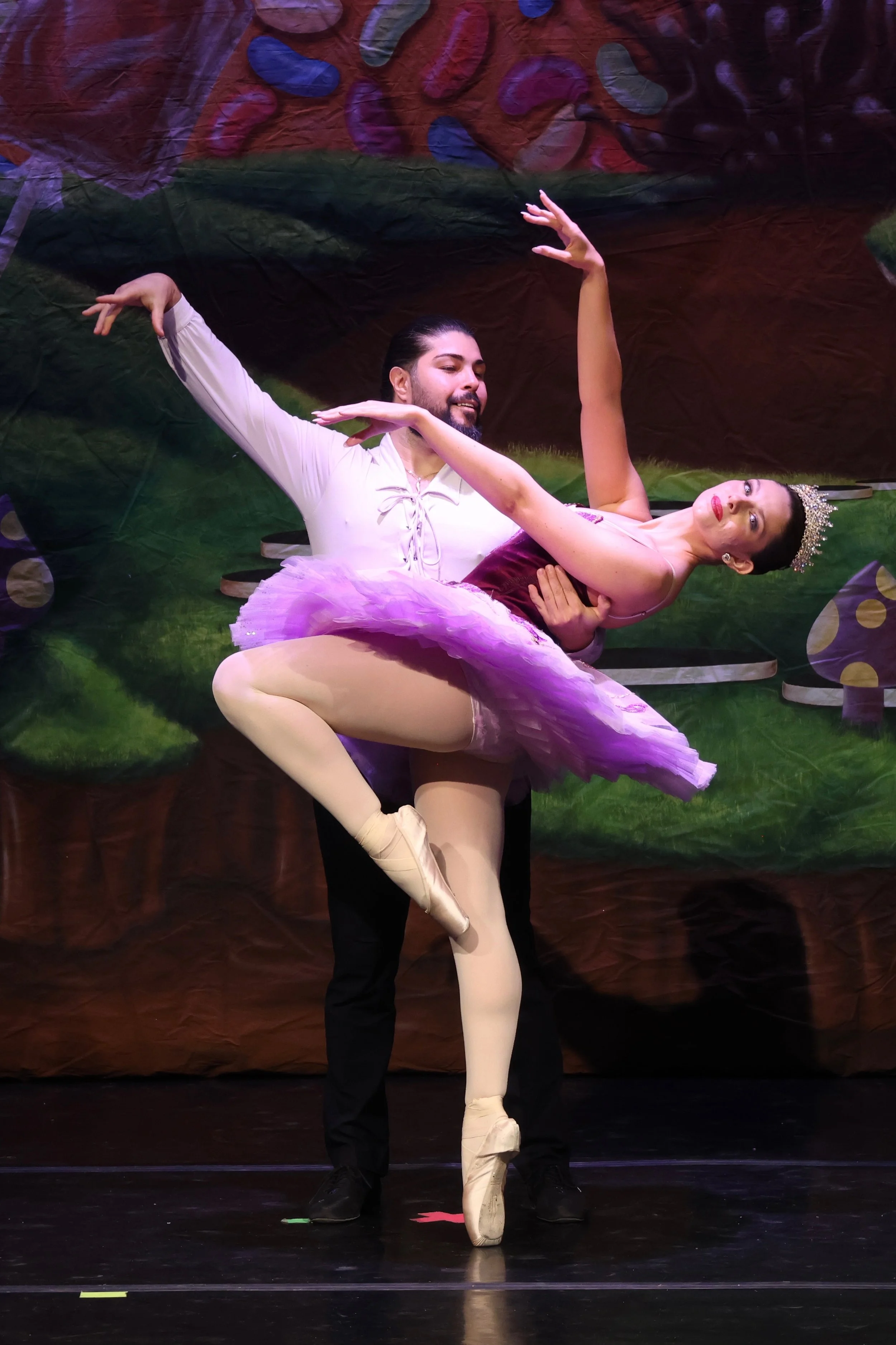 Ballet dancers performing on stage with a male dancer supporting a female dancer in a pose, background decorated with colorful abstract patterns.
