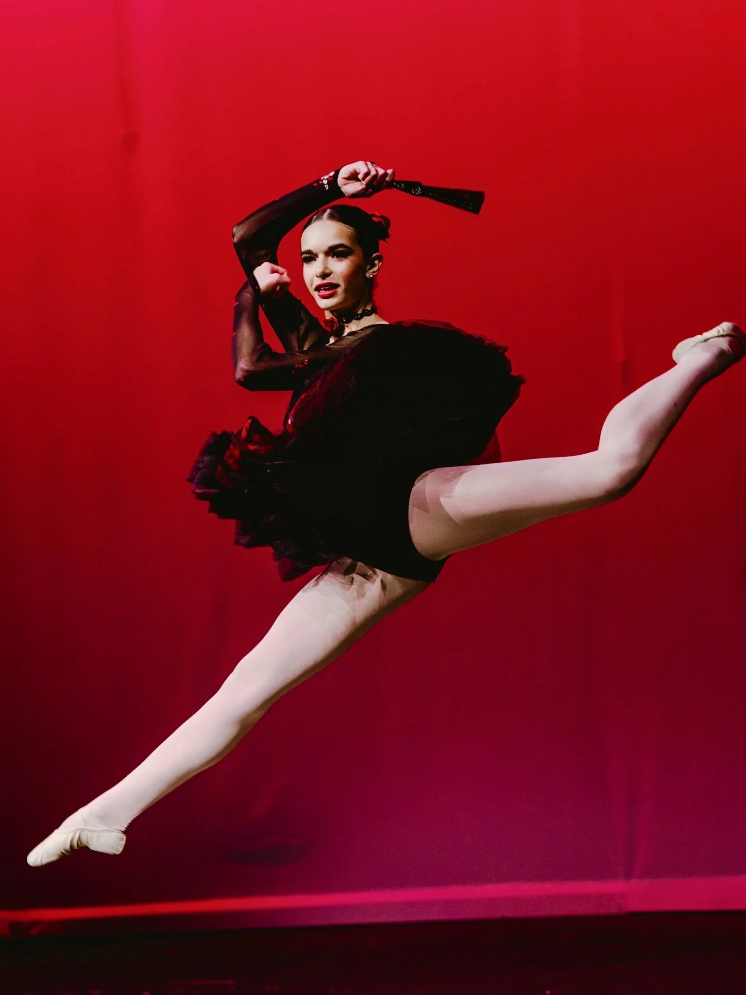 A ballet dancer performing a split leap against a vibrant red background, dressed in a black and beige costume with pink pointe shoes.