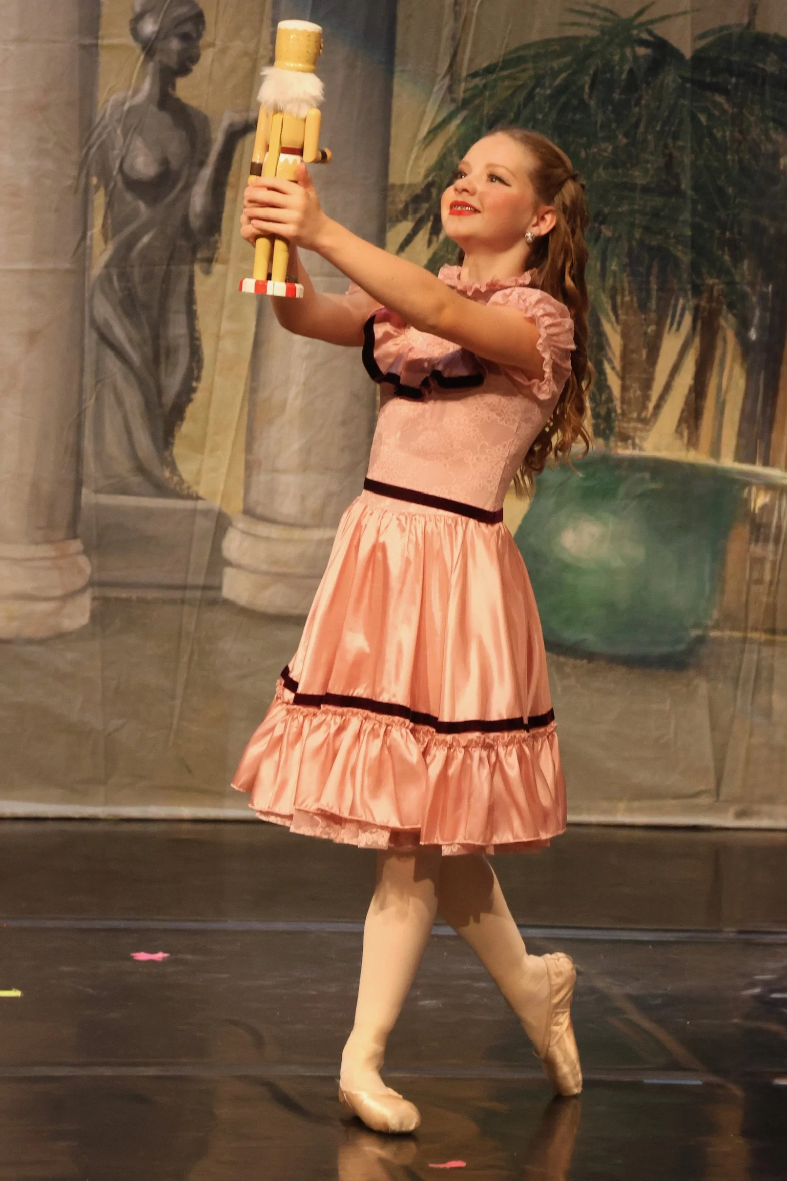 A young woman in a pink ballet dress and pointe shoes dancing on stage, holding a toy soldier in her right hand, with a painted backdrop of a statue and trees behind her.