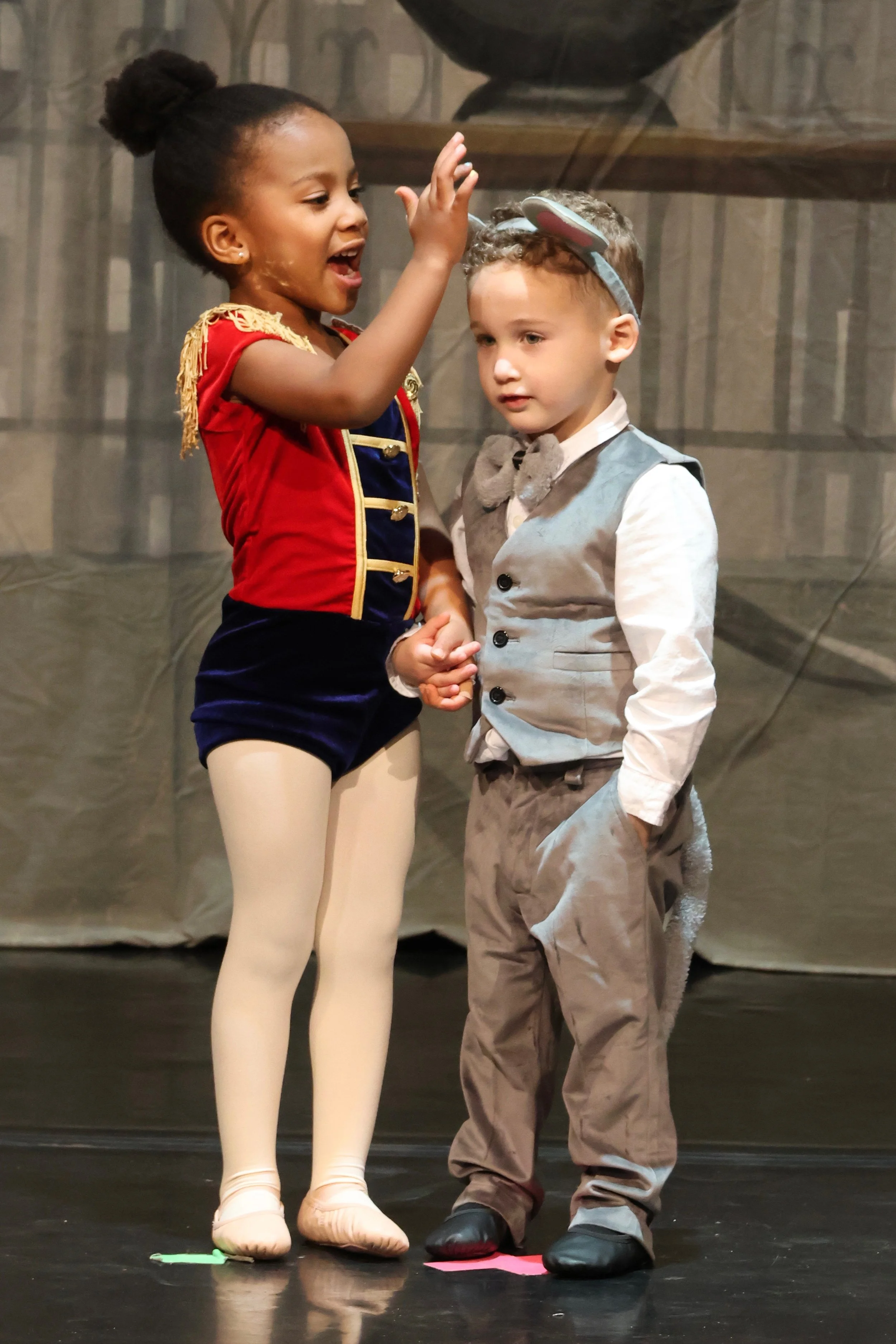 Two children, a girl dressed as a ballerina and a boy dressed as a circus clown, holding hands on stage during a performance or rehearsal.