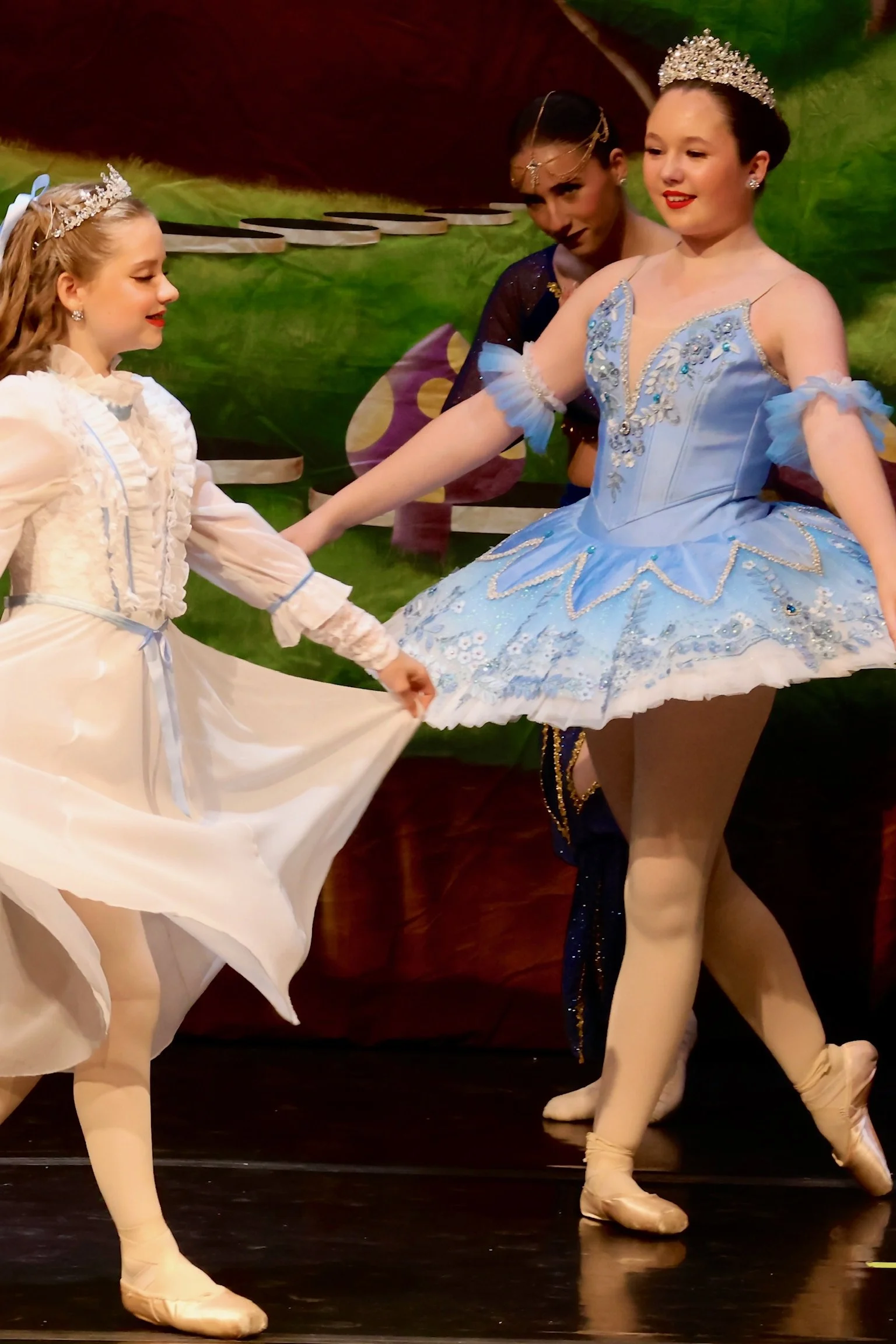 Ballet dancers on stage, two girls in cream-colored tutus and one girl in a blue tutu, all wearing tiaras and ballet shoes, performing a dance.