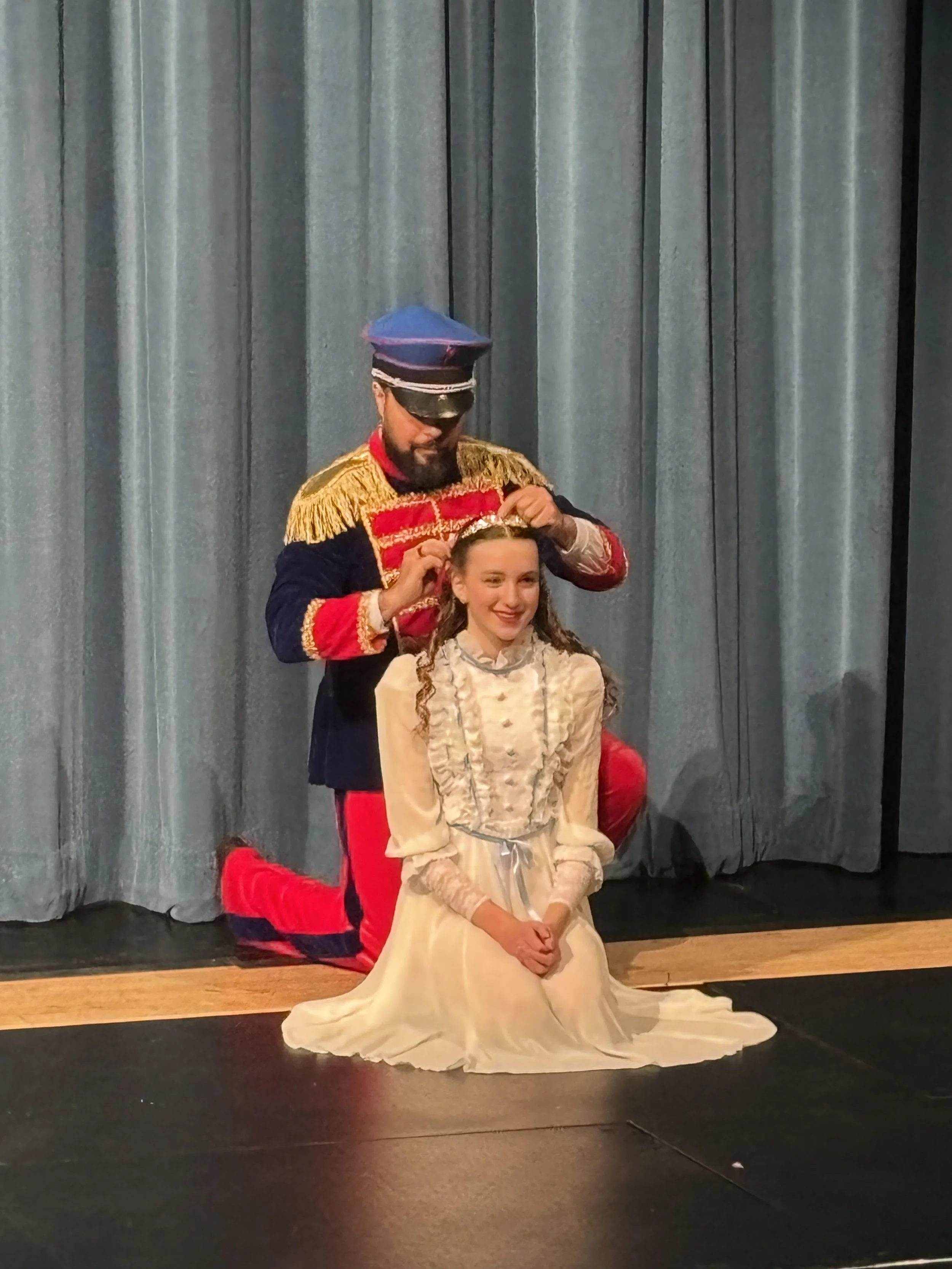 A man dressed as a marching band leader is kneeling behind a young girl in a beige dress, helping her with a costume or accessory on her head. They are on stage with gray curtains in the background.