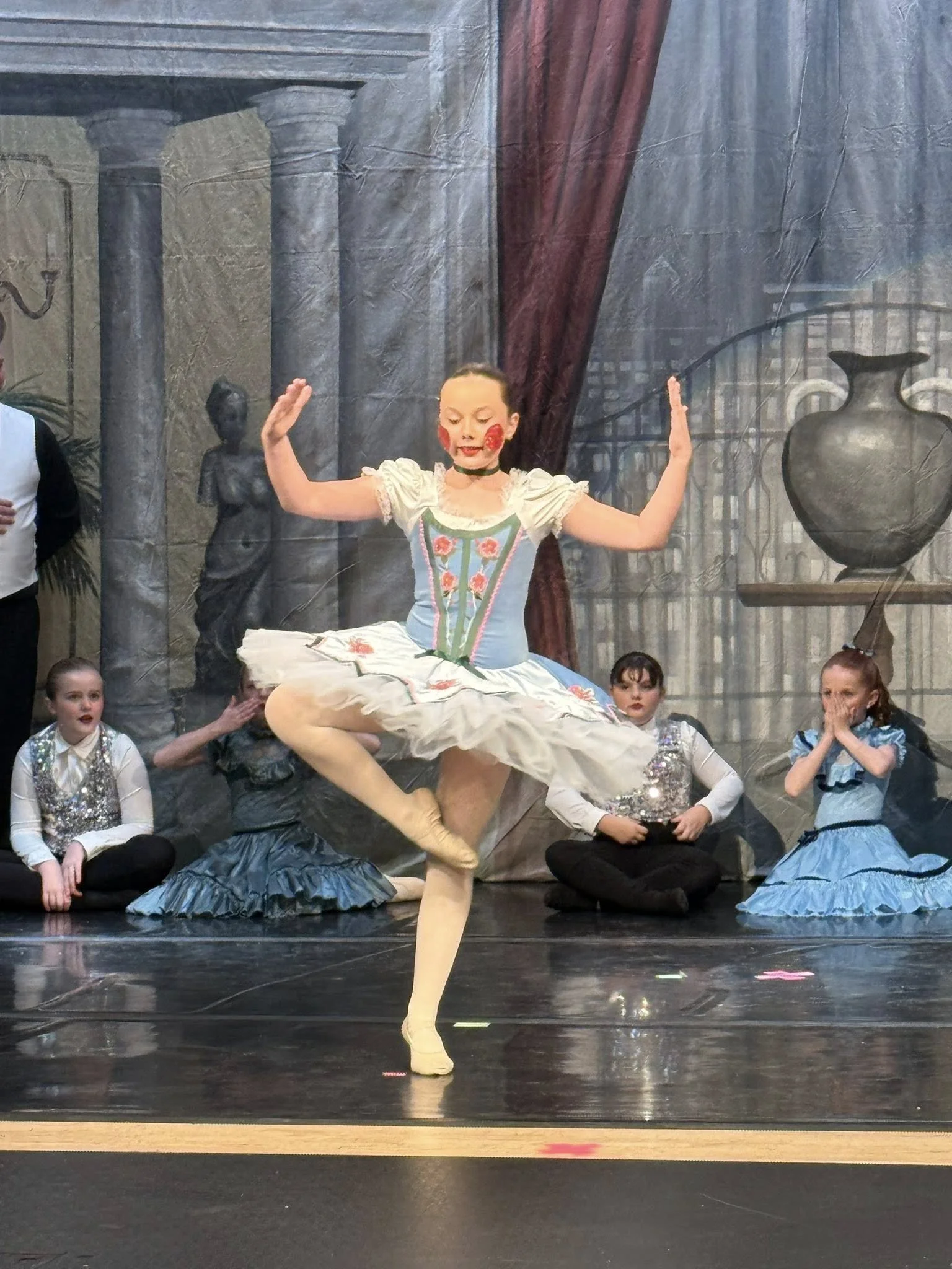 Ballet dancer in a tutu performing on stage during a performance, with children and actors in costumes watching in the background.