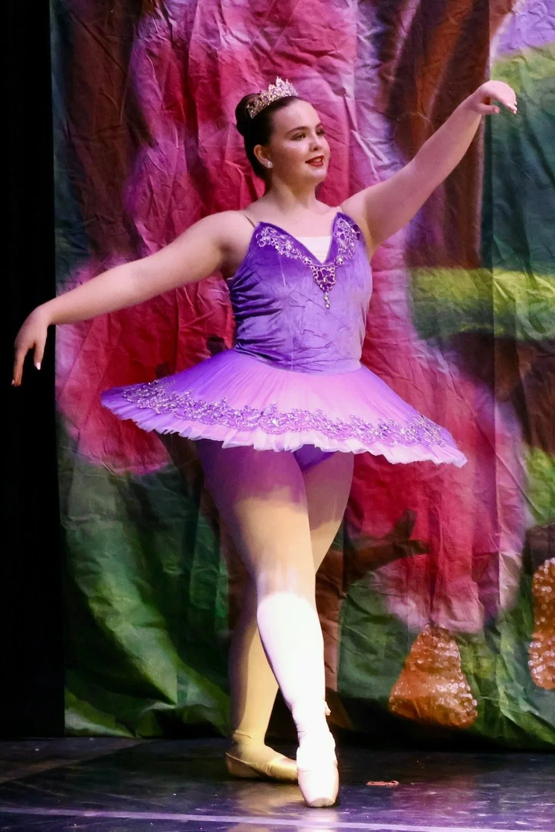 Ballet dancer in a purple tutu and tiara on stage, mid-pose, with a colorful backdrop.