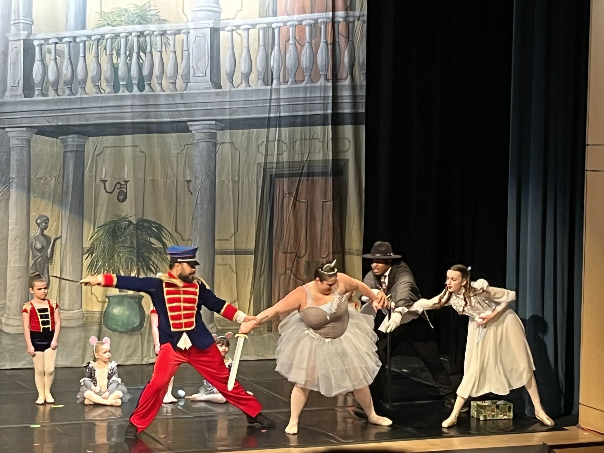 The stage scene depicts a theatrical performance with actors in period costumes, some sitting and some standing, engaged in a dramatic scene with a backdrop resembling a classical European balcony and interior decor.