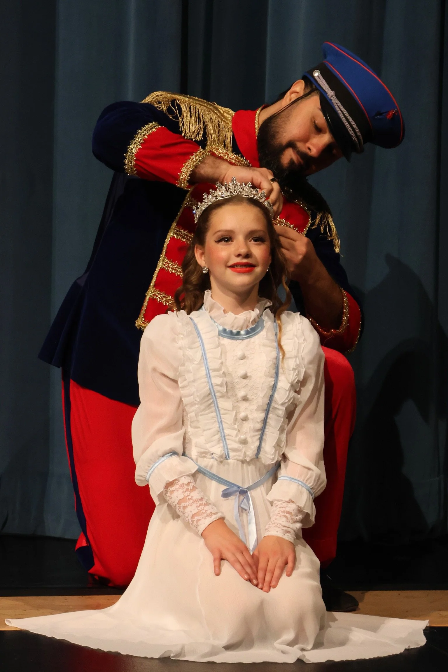 A young girl in a white dress with lace details and a light blue ribbon sash, seated on the floor, being crowned with a tiara by a man in a ceremonial uniform with red and navy blue and gold accents.