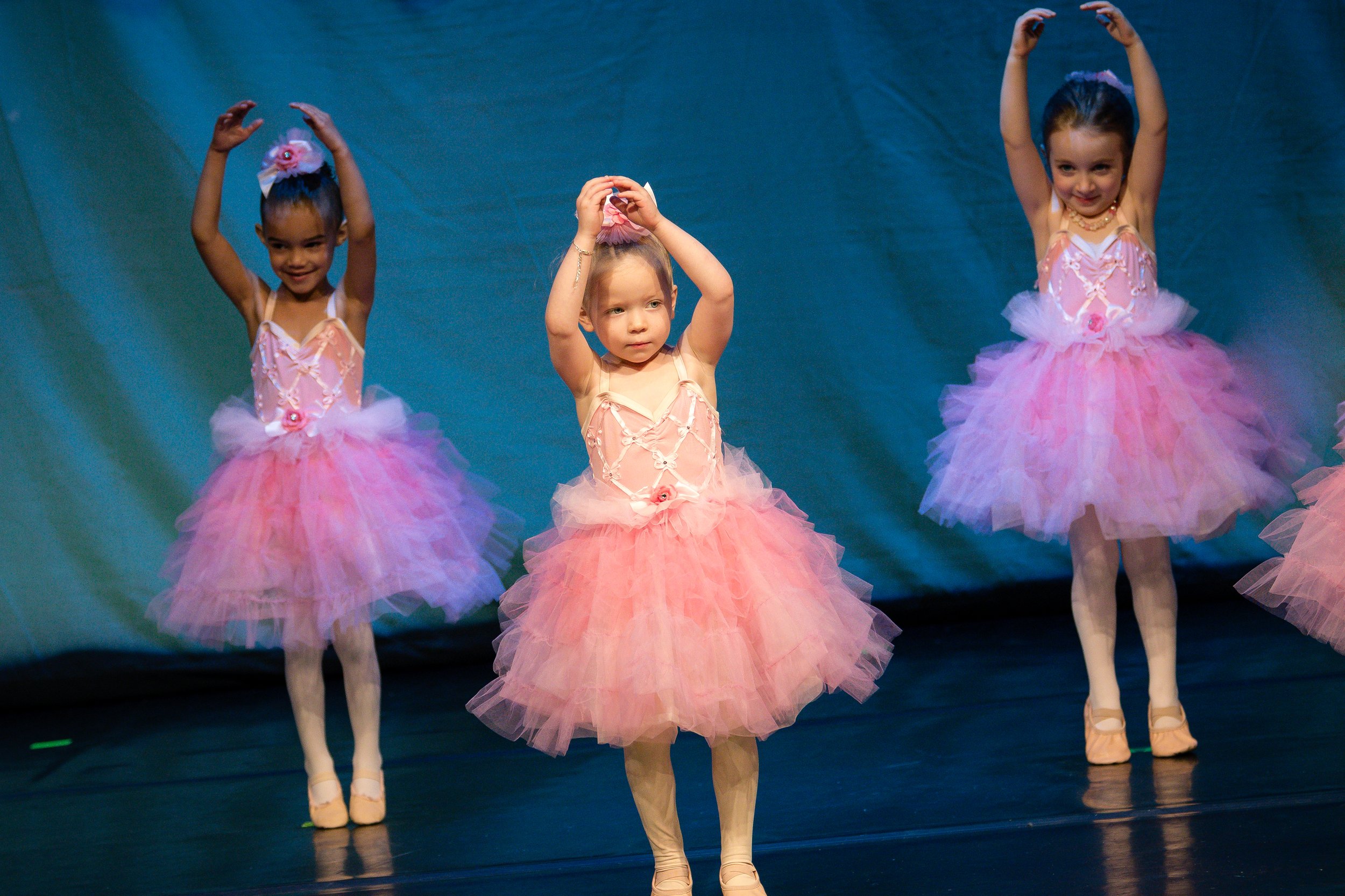 Six young girls in pink tutus holding hands on stage during a dance performance.