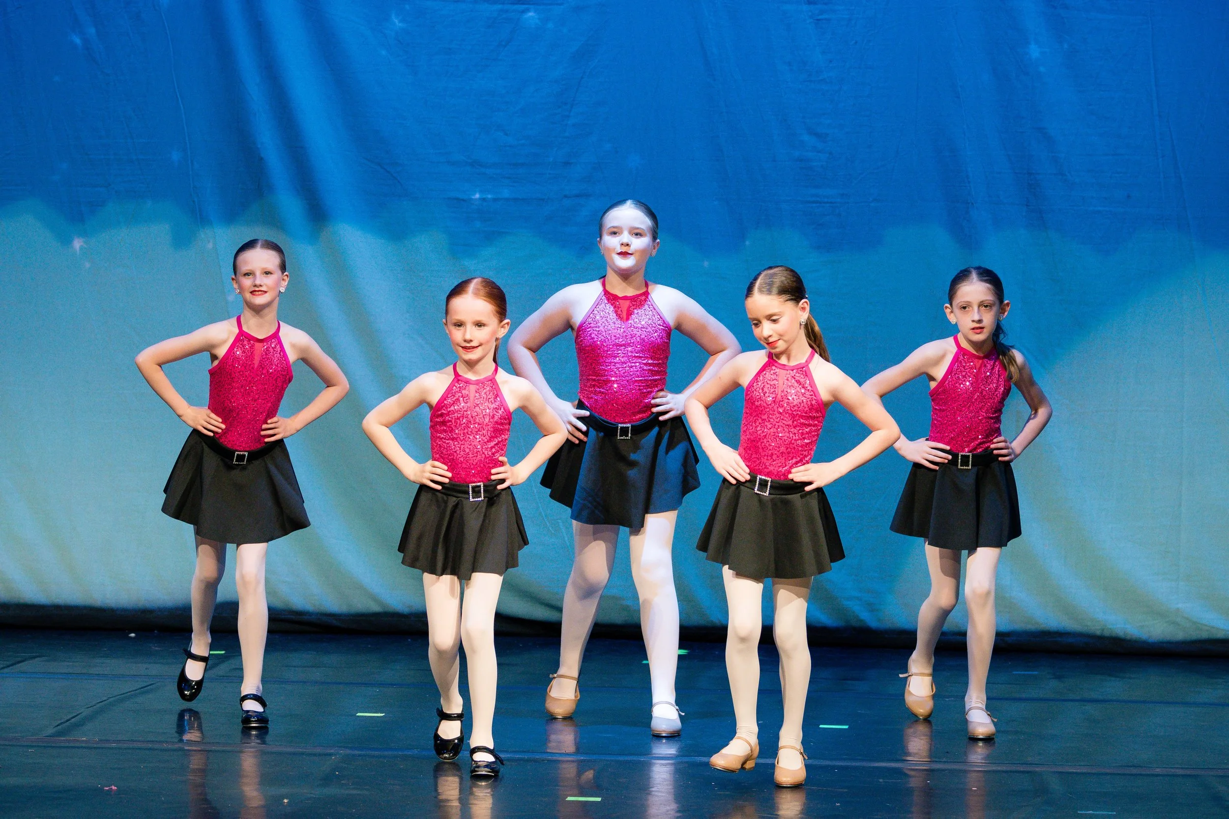 Five young girls in pink sequin tops, black skirts, and tights performing a dance on stage with a blue curtain background.