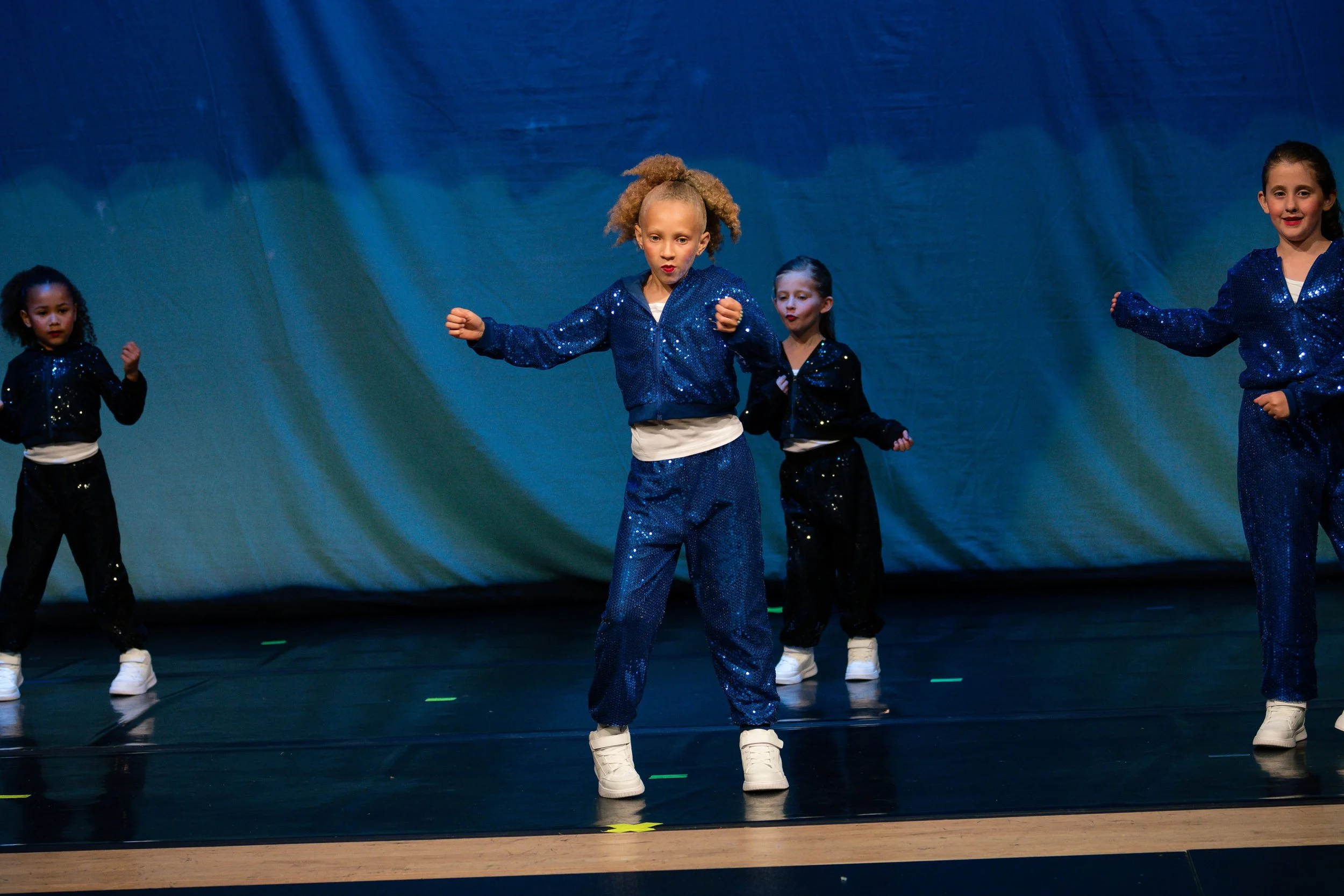Four young girls performing a dance on stage in matching blue and black sequined outfits.