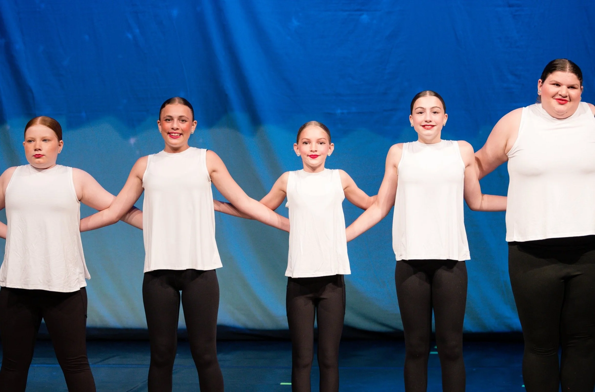 Group of five young female dancers in white tops and black leggings standing in a line holding hands on stage, blue curtain backdrop.