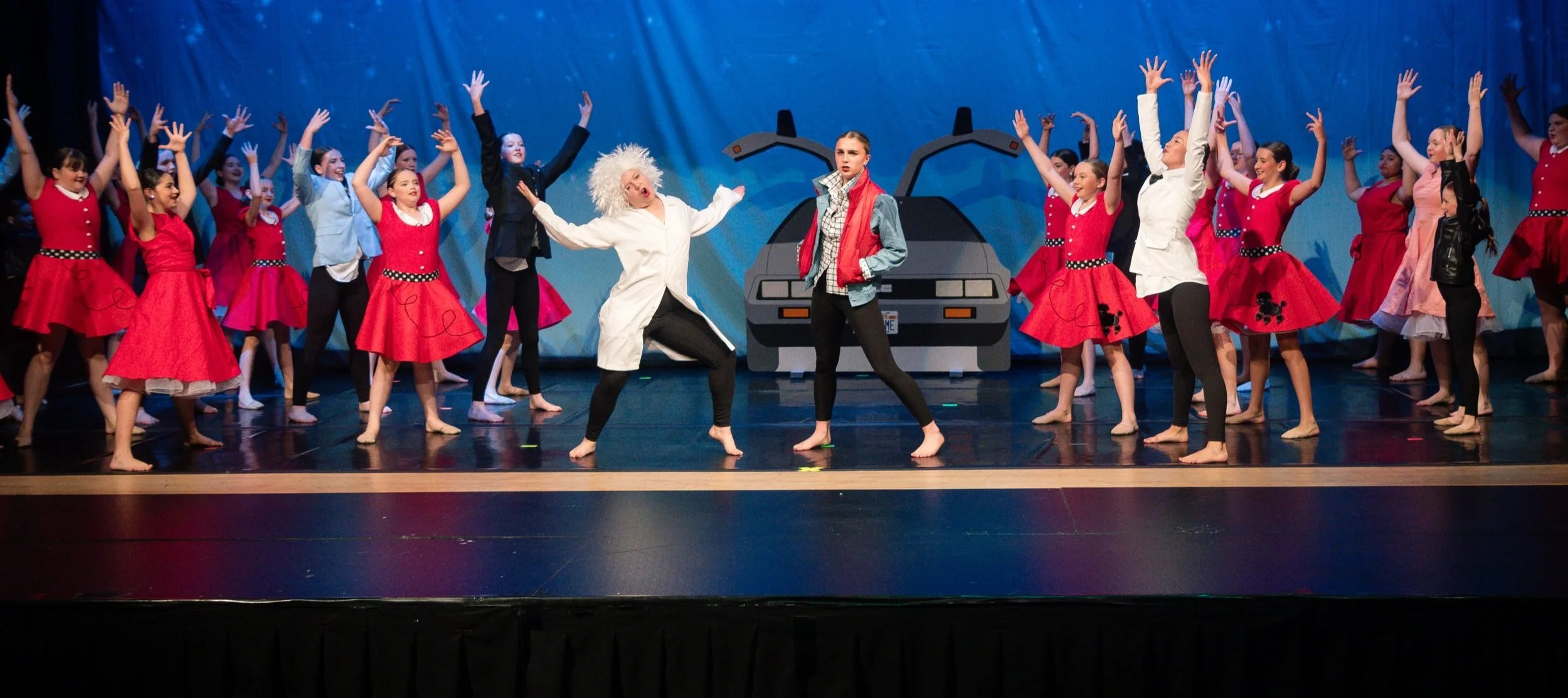 Scene from a theatrical dance performance featuring children in costumes, with some dressed as characters from 'Back to the Future,' on stage with a futuristic backdrop and a DeLorean car.