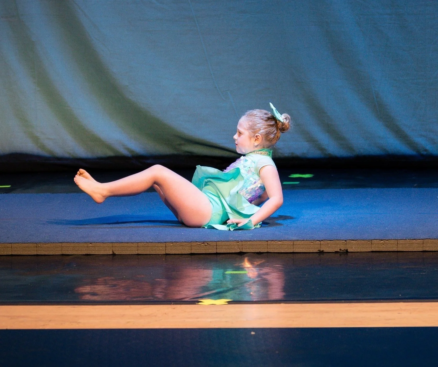 Young girl in a green costume sitting on a stage with her legs extended and leaning back, with a blue backdrop.