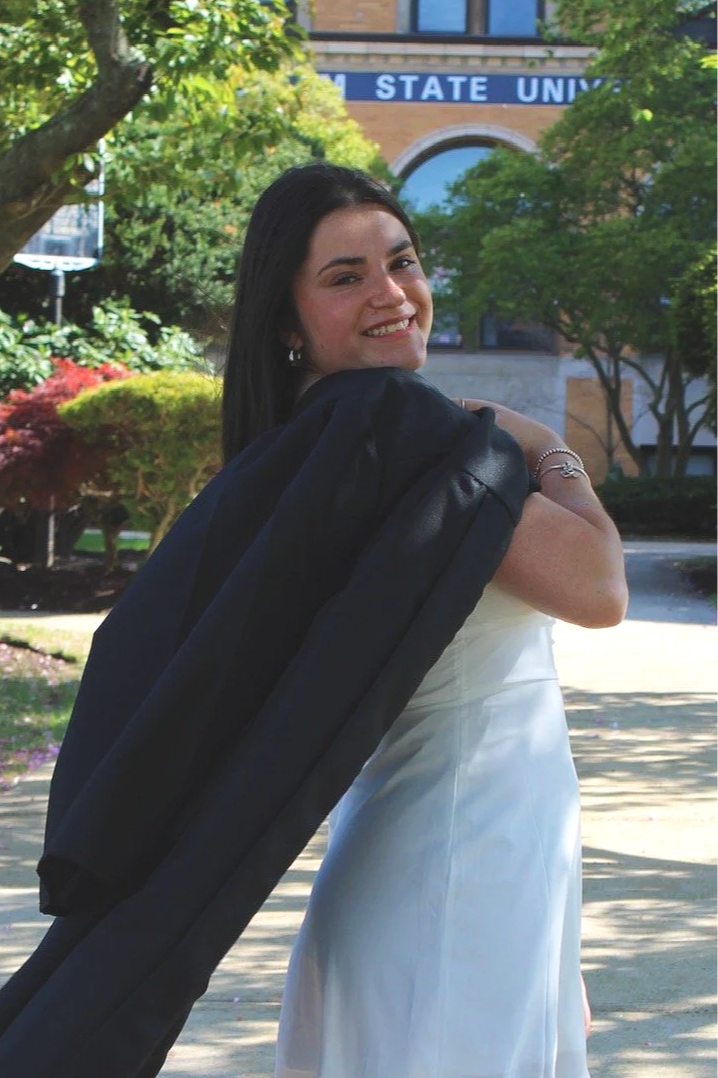 Young woman in a white dress smiling, carrying a black graduation gown over her shoulder on a university campus.