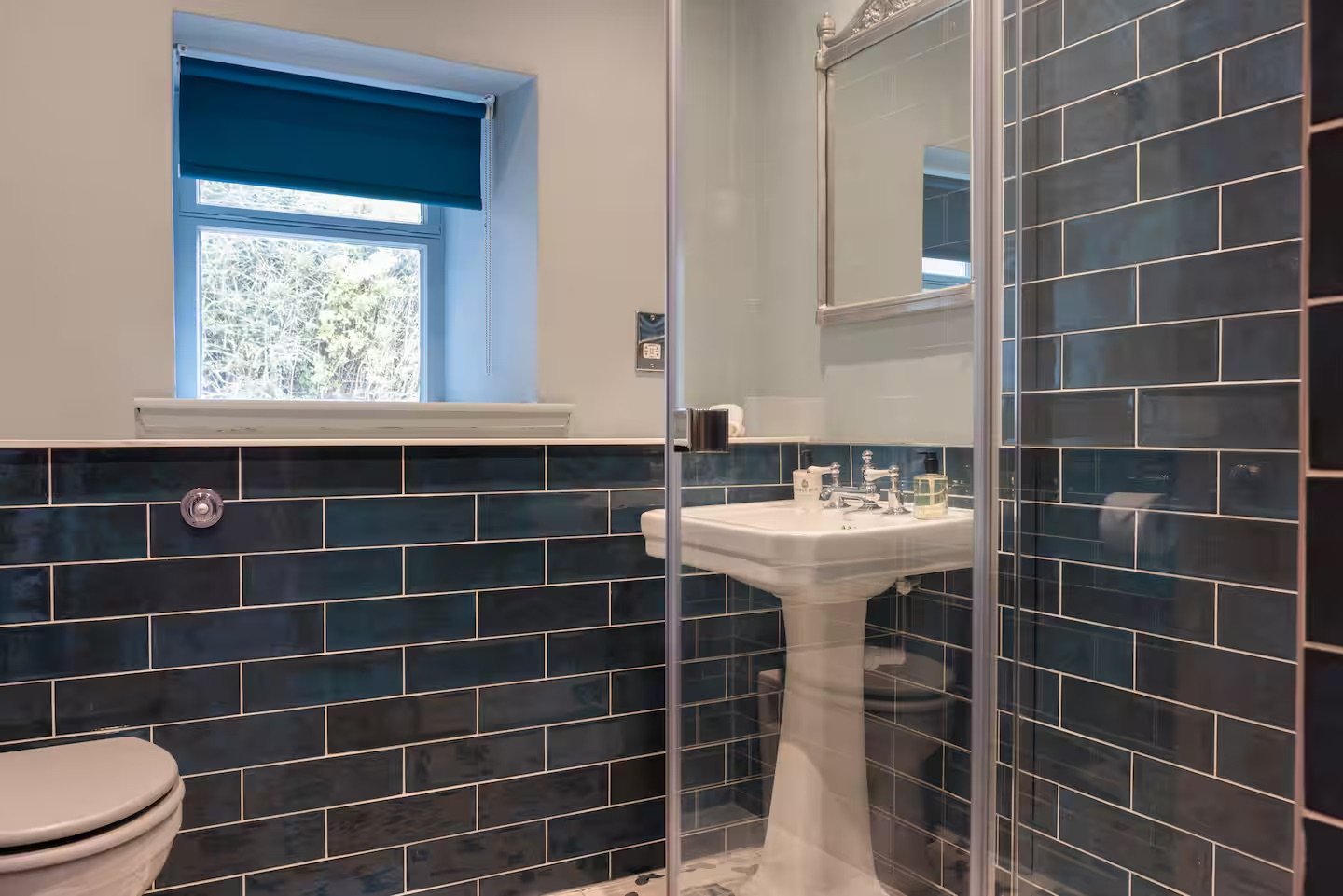 A small bathroom with dark blue subway tile walls, a pedestal sink with a mirror above, a glass-enclosed shower, a window with a blue shade, and a toilet in the corner.