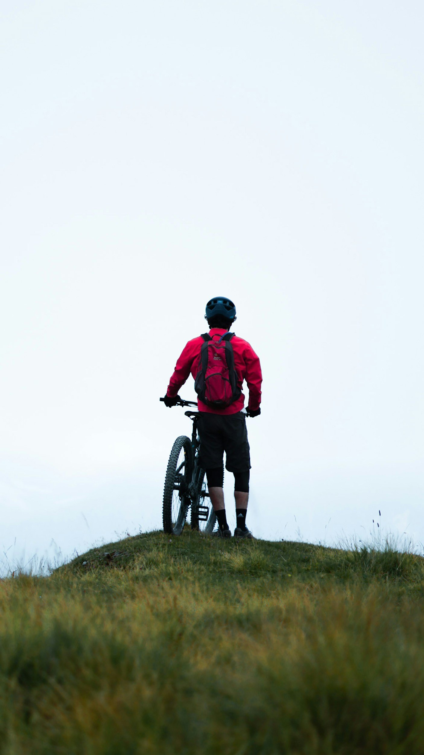 A person dressed in a red jacket and black shorts, wearing a helmet and backpack, standing on a small grassy hill and holding a mountain bike, looking into a foggy, overcast landscape.