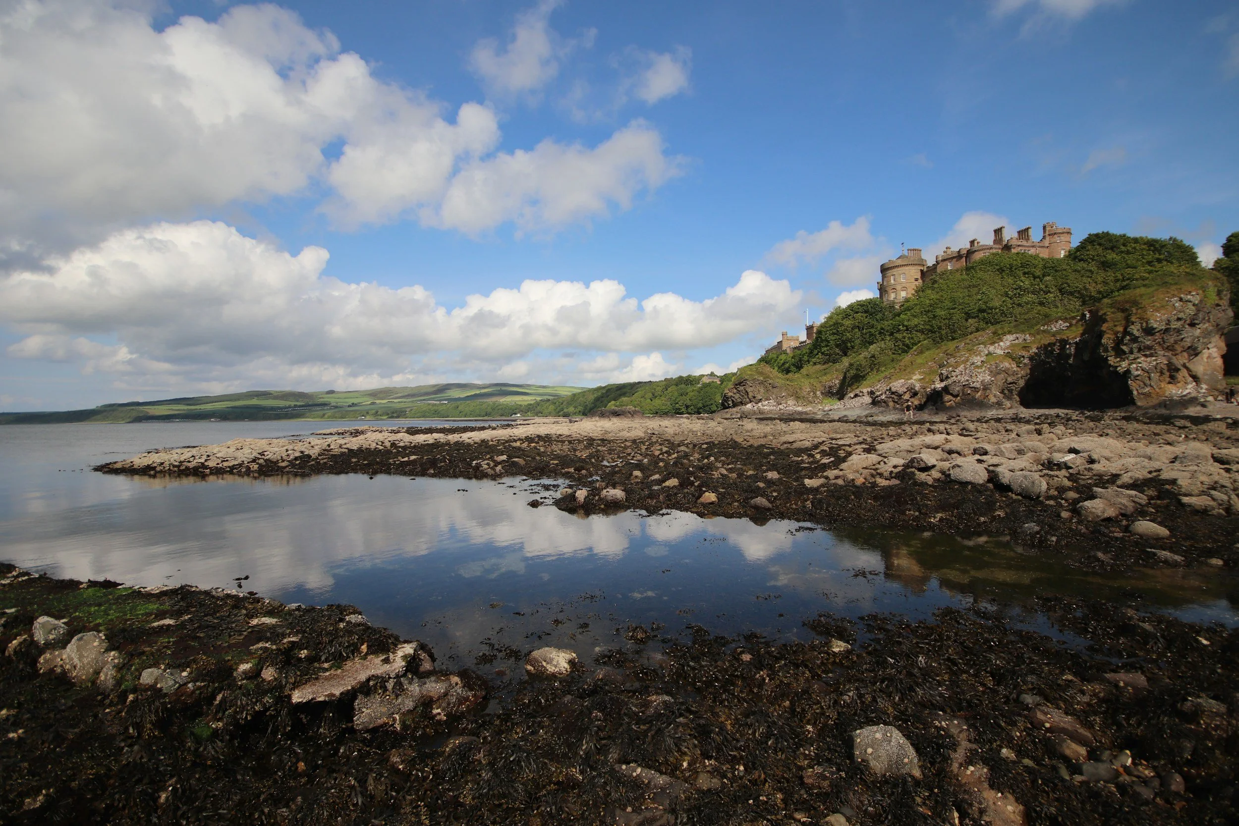 A castle on a hill overlooking a body of water with rocks and greenery in the foreground, under a partly cloudy sky.