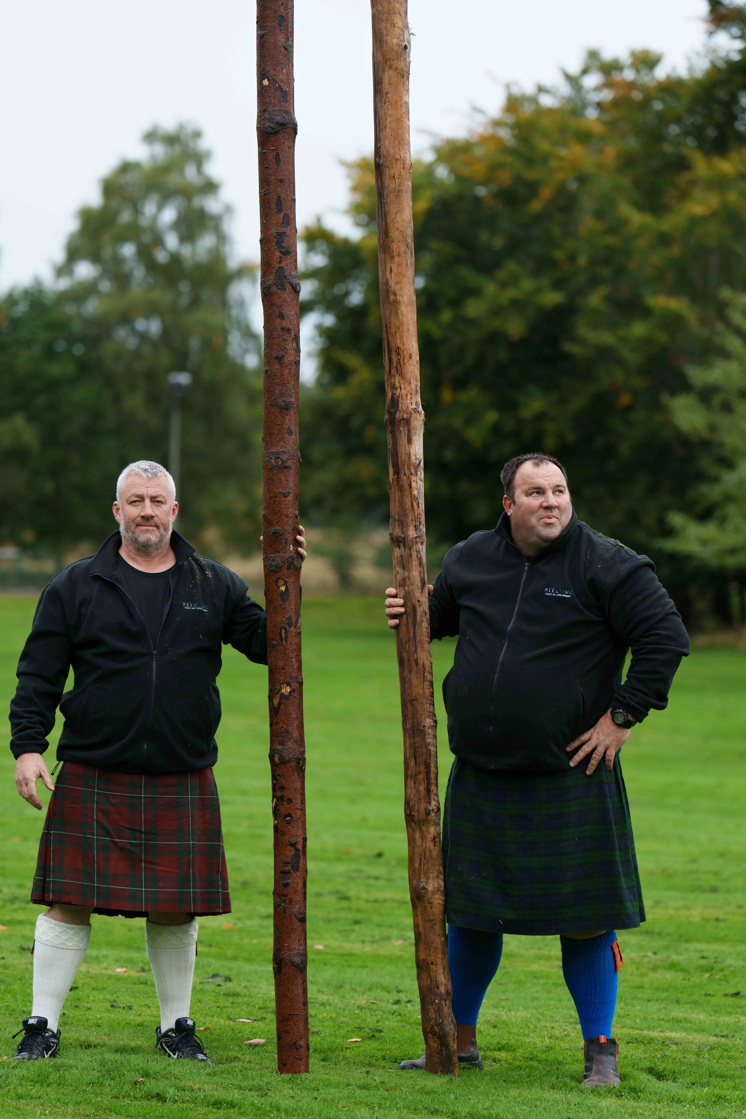 Two men wearing traditional Scottish kilts, holding wooden poles, standing outdoors on a grassy field with trees in the background.