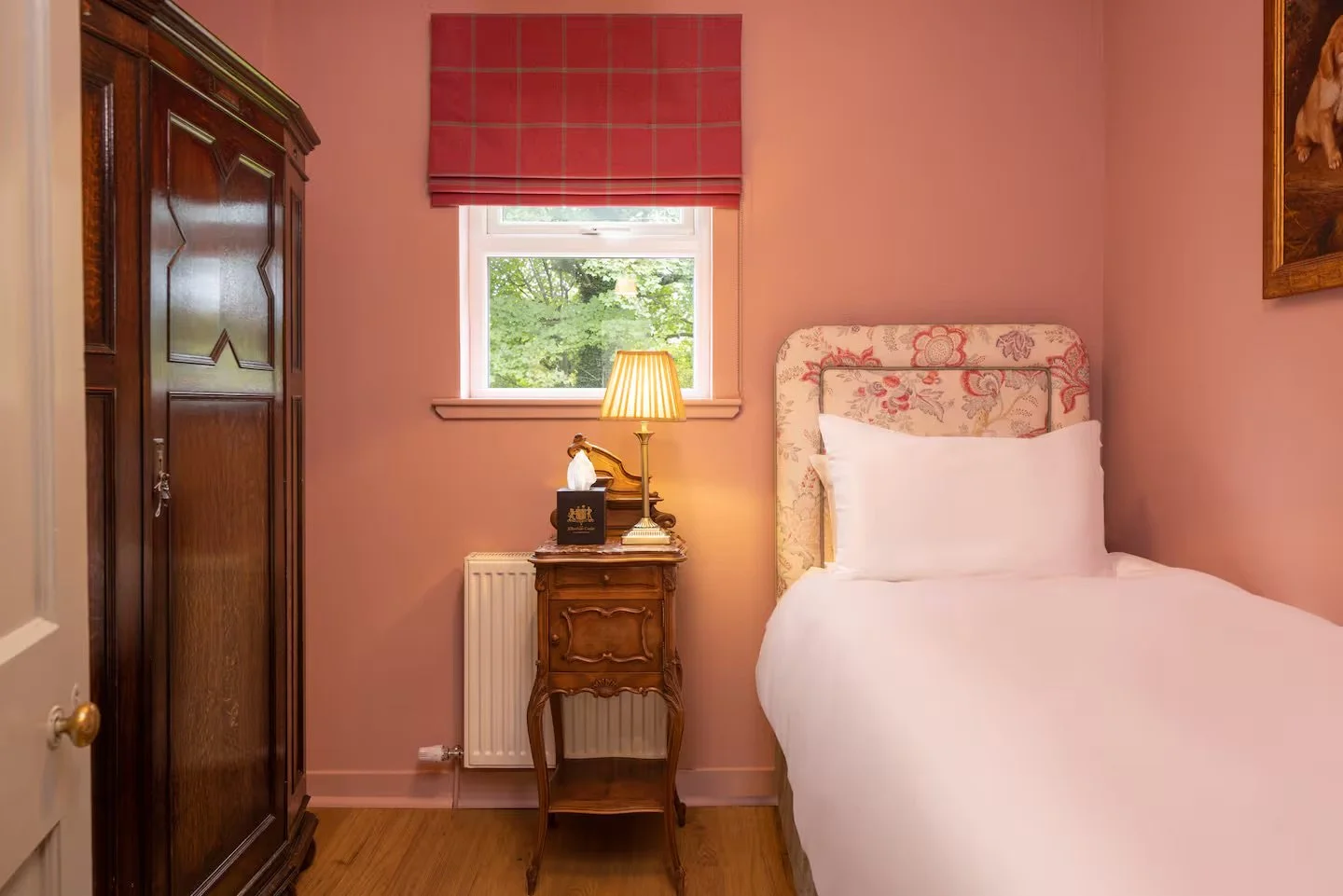 A cozy bedroom corner with a single bed, floral headboard, pink painted walls, and a window with a red accordion shade showing green trees outside. There is a wooden wardrobe, a small wooden bedside table with a lamp, tissue box, and a painting hanging on the wall.