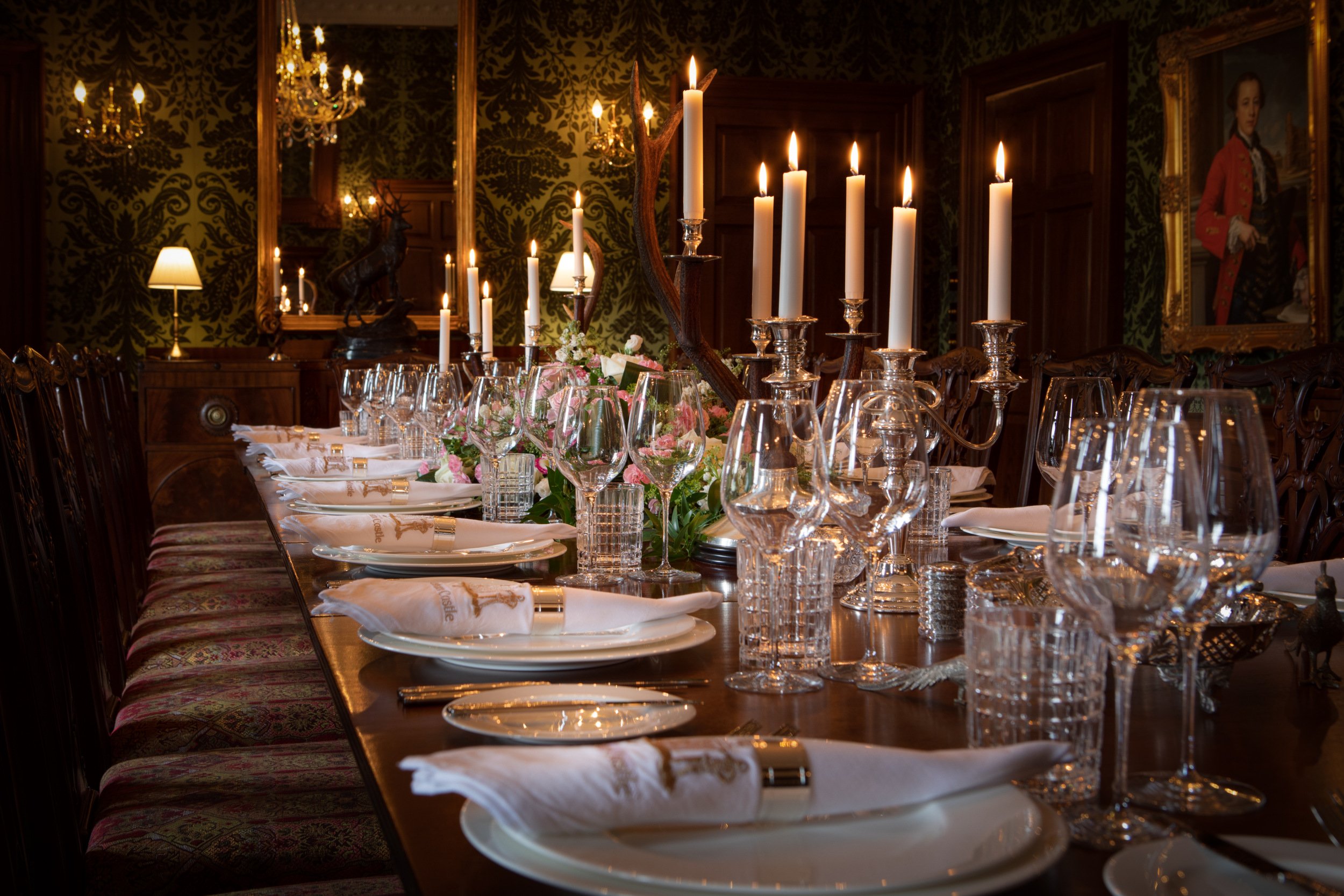 Elegant dining table set with wine glasses, silver candlesticks with lit candles, floral centerpiece, and neatly arranged plates and napkins in a richly decorated room with dark wallpaper, chandeliers, paintings, and warm lighting.