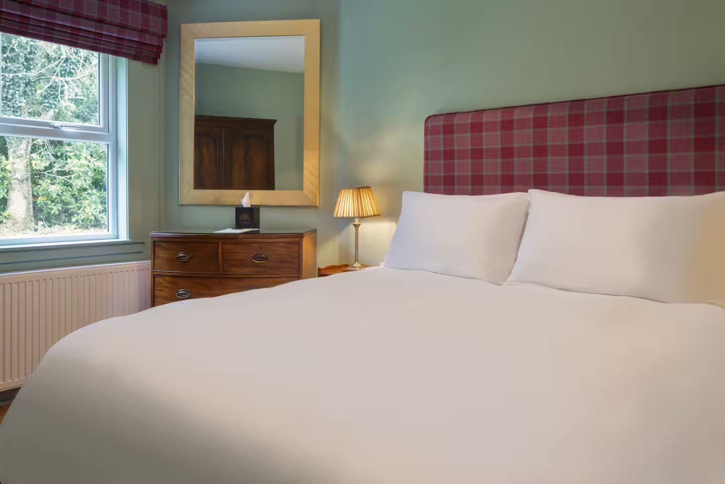 A neatly made bed with white pillows and a checked red and green headboard in a cozy hotel room. A window with blinds and a view of trees outside, a wooden dresser with a mirror above, and a small lamp on the bedside table are visible.