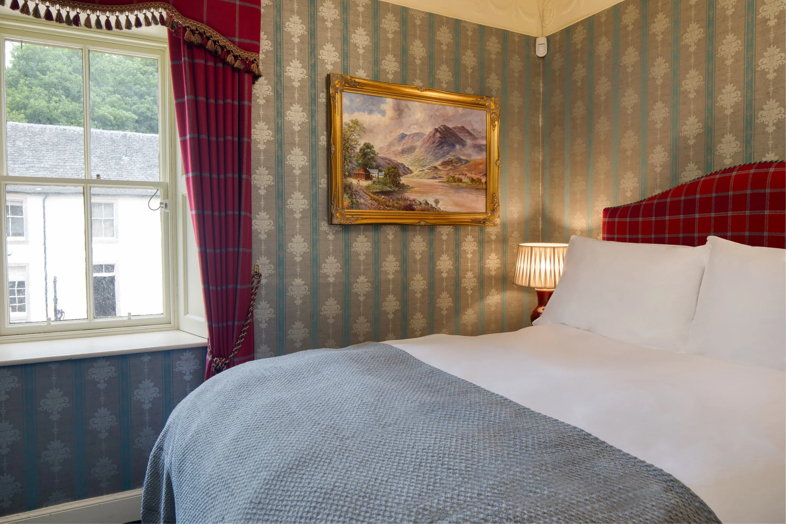 A bedroom with wallpaper, a window with red curtains, a landscape painting in a golden frame, a bedside lamp, and part of a bed with white pillows and a gray blanket.