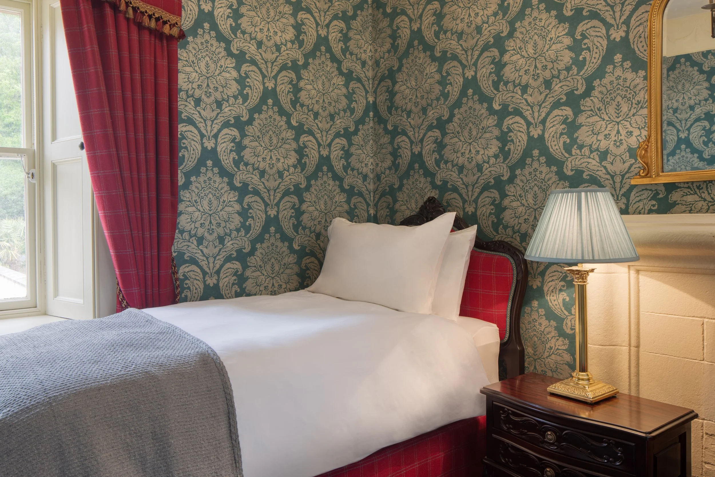 A bedroom with a vintage floral damask wallpaper in blue and beige, a window with red plaid curtains, a bed with white bedding and pillows, a bedside table with a blue lampshade and a gold-framed mirror on the wall.