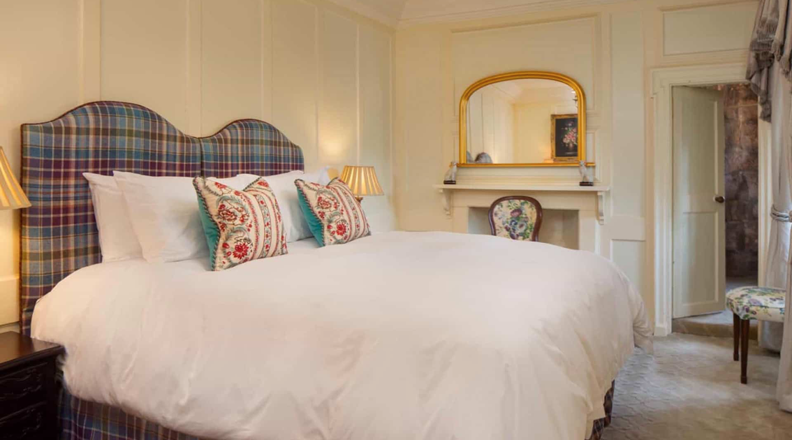 A cozy bedroom with a large bed featuring a tartan upholstered headboard, decorative pillows, and two lamps on bedside tables. There is a mirror above a small fireplace, and a floral chair is positioned near the doorway, with curtains on the window.