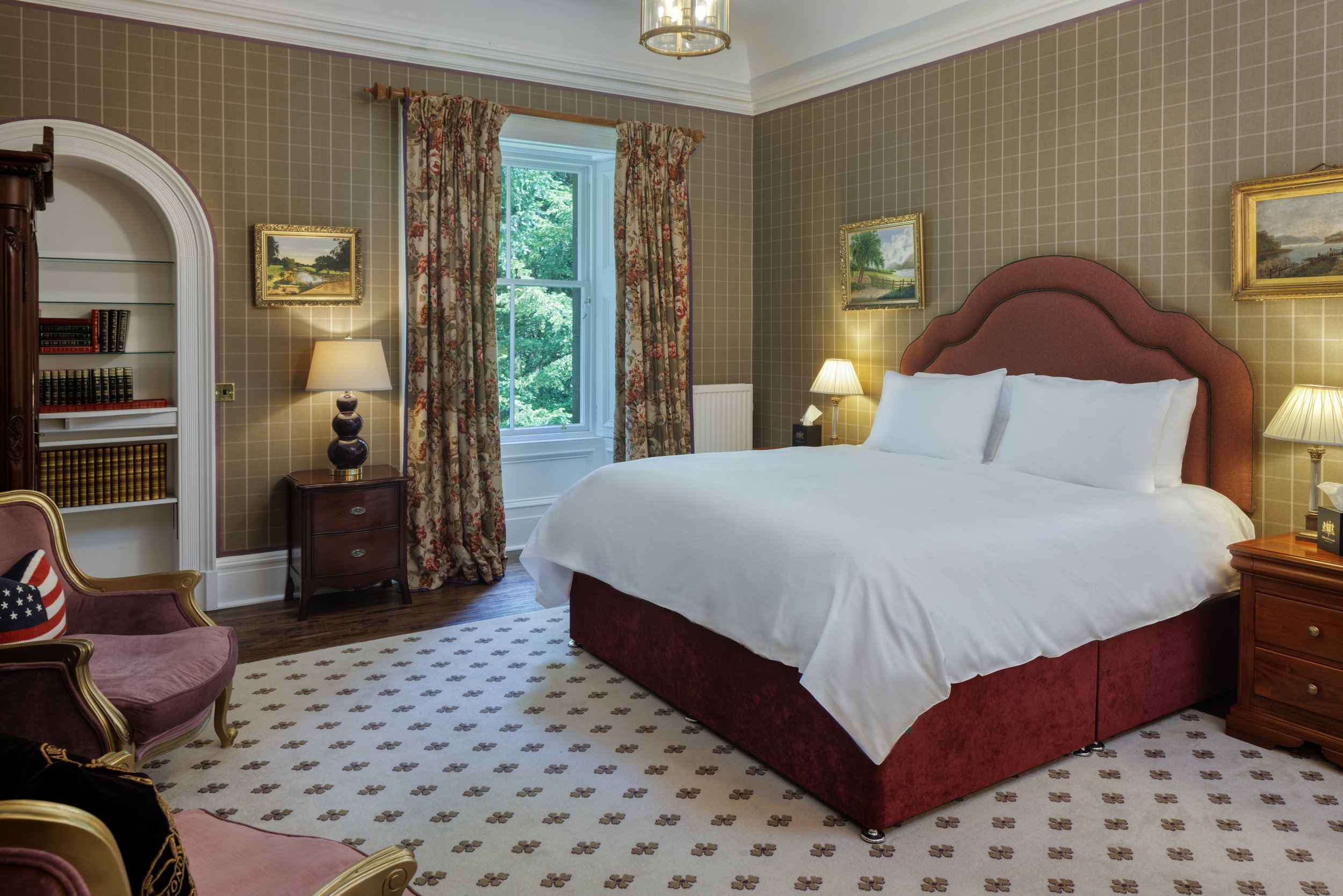 A bedroom with a large bed with a pinkish-red headboard, white bedding, and pillows. There is a window with floral curtains, two table lamps on wooden nightstands, a bookshelf filled with books, a pink armchair with an American flag pillow, and framed landscape paintings on the beige plaid wallpapered walls.