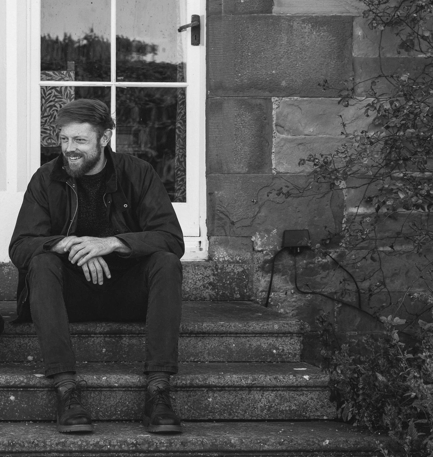 A man with a beard and short hair sitting on stone steps in front of a building with a window. He is smiling and wearing a dark jacket, dark pants, and boots.