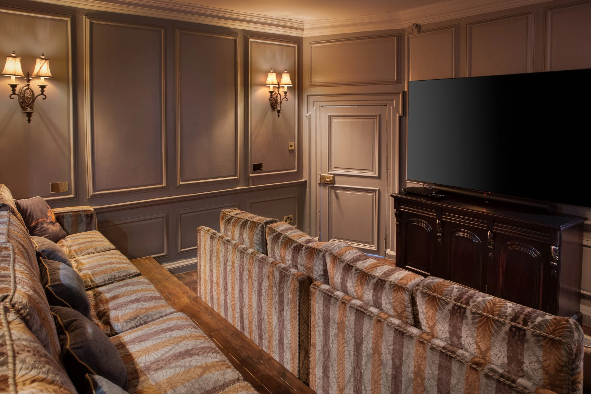 Living room with beige paneled walls, wall sconces, and a large flat-screen TV on a dark wooden stand. Multiple upholstered sofas with patterned fabric are arranged in the room.
