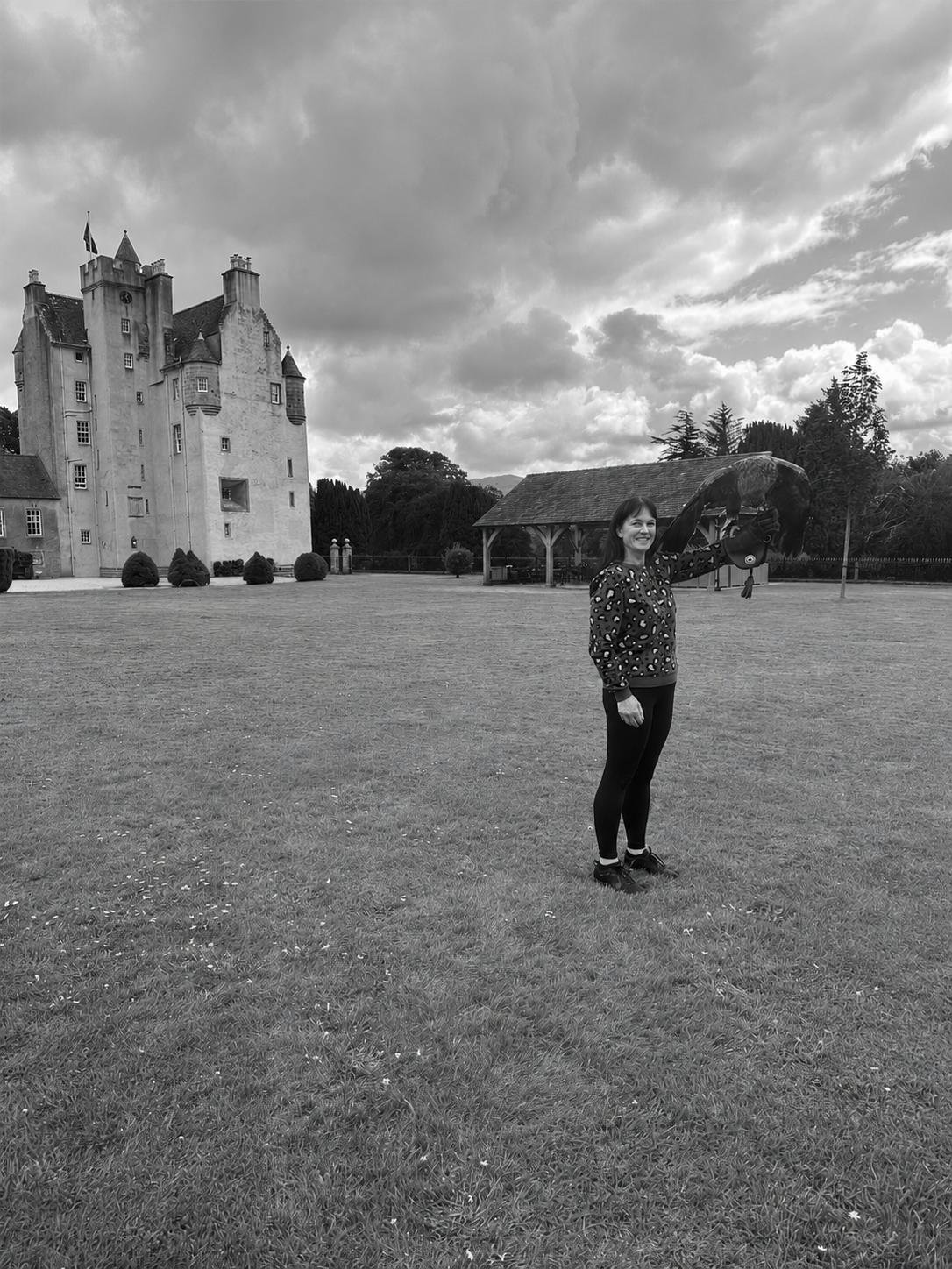 A woman standing on a lawn with a castle in the background, holding a headless horse statue, against a cloudy sky.