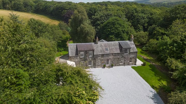 A large stone house surrounded by lush green trees and grass, with a gravel driveway in front.