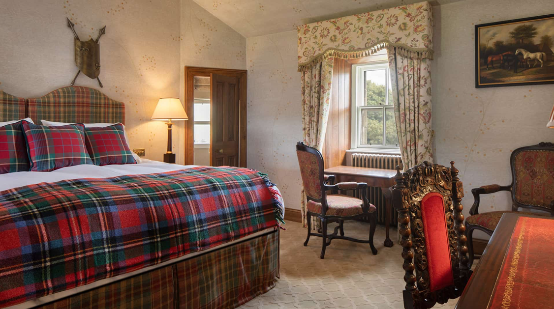 A cozy bedroom with a plaid bedspread, vintage wooden furniture, a window with floral curtains, and a painting of horses on the wall.