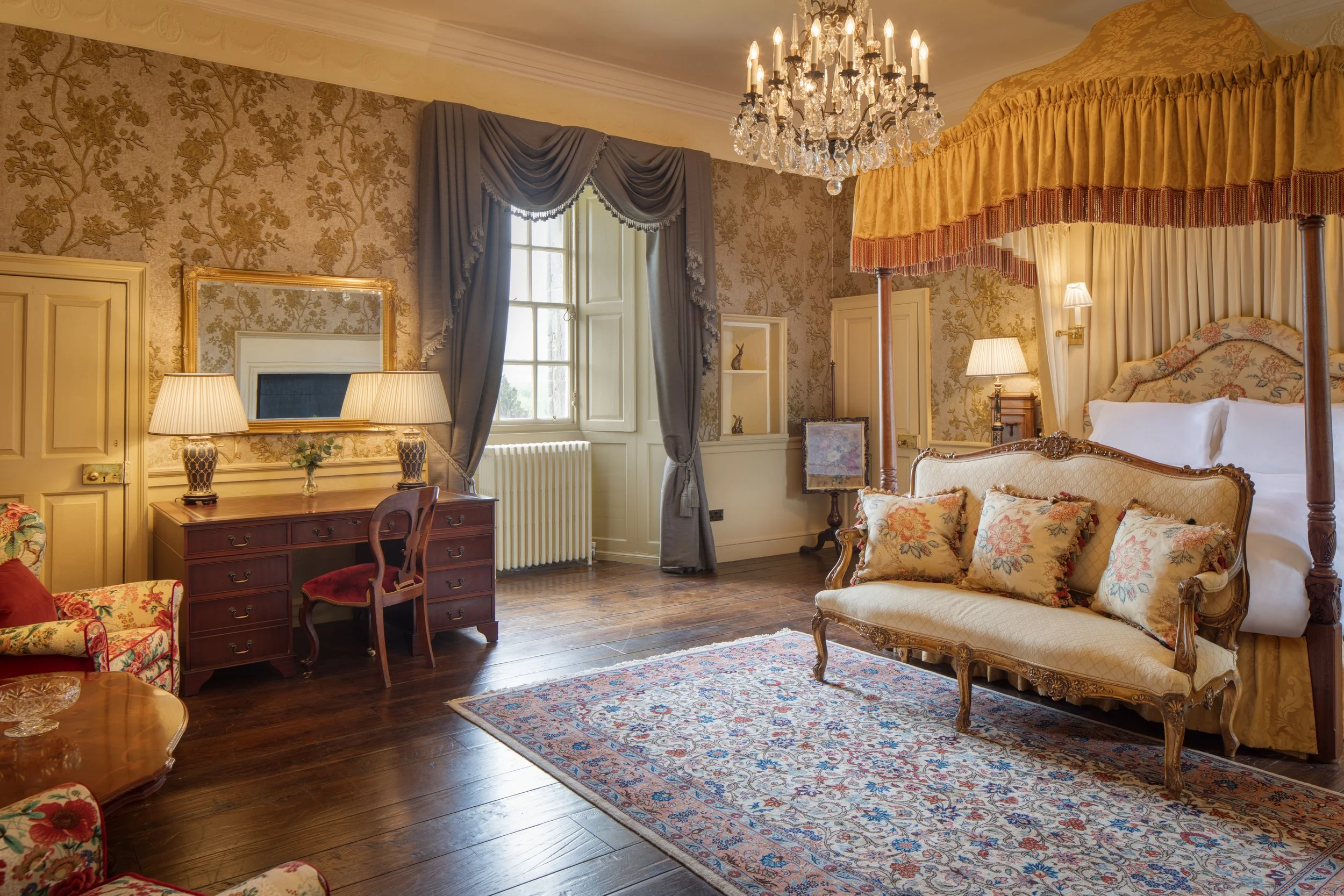 A luxurious vintage bedroom featuring a four-poster bed with floral and gold upholstery, a crystal chandelier, a wooden dresser with table lamps, floral wallpaper, and a large ornate mirror.