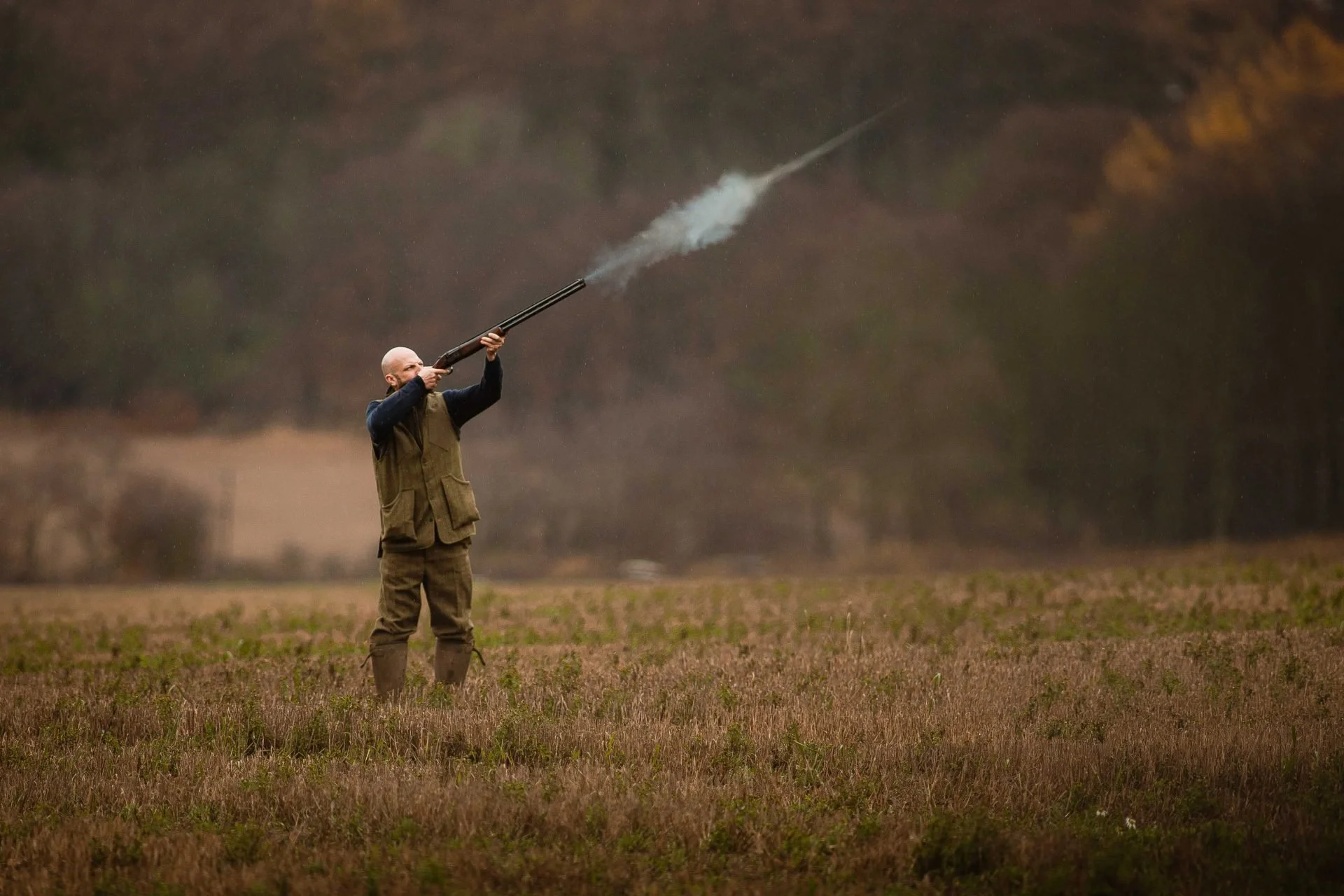 A man outdoors in a field, wearing camouflage boots and a brown vest over dark clothing, aiming a double-barrel shotgun as smoke and a shot are emitted from it.