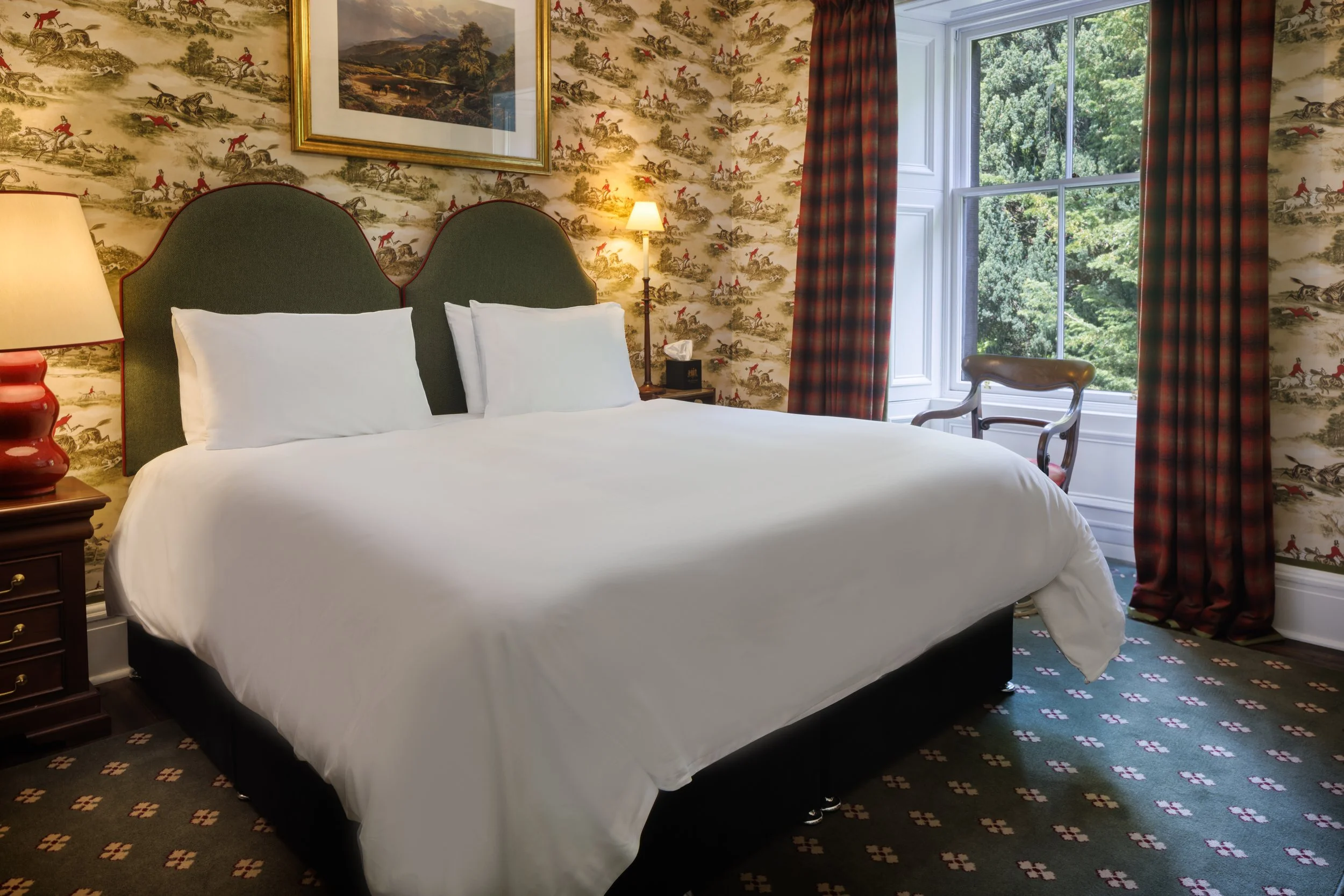 A hotel room with a large bed, green upholstered headboard, white bedding and pillows, two lamps, floral wallpaper, a window with red plaid curtains, and a wooden chair outside the window.